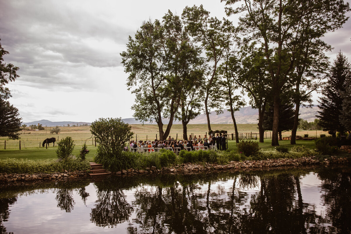 Wedding ceremony taking place beneath tall trees beside a pond on a ranch, with a grazing horse in the background and mountains in the distance under a cloudy sky.