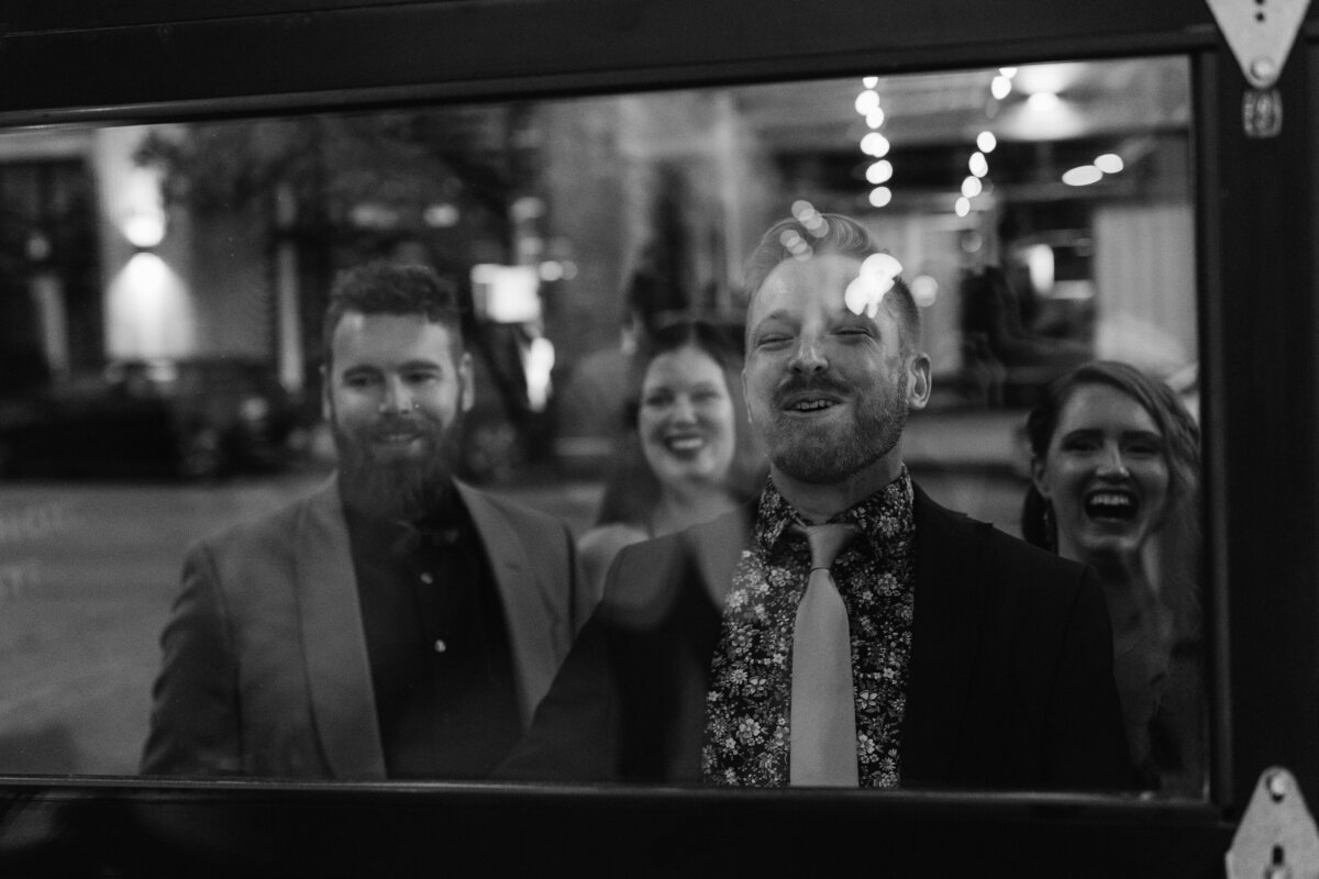 Black and white photo of wedding guests laughing and enjoying themselves, seen through a window with city lights in the background