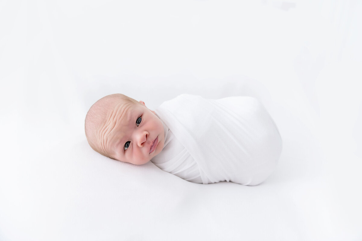 An awake newborn baby lays on a white bed wrapped tightly in a white swaddle