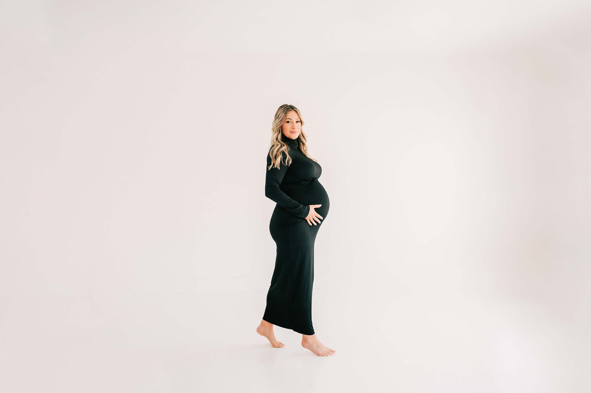 matenrity photo of pregnant mom in black dress walking in white photography studio in Springfield MO