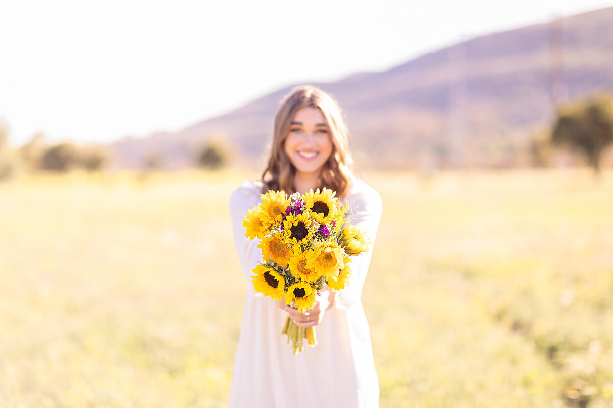 A person in a white dress stands in a sunlit field, smiling and holding a bouquet of yellow sunflowers. The background is a blurred landscape with a hill and trees.