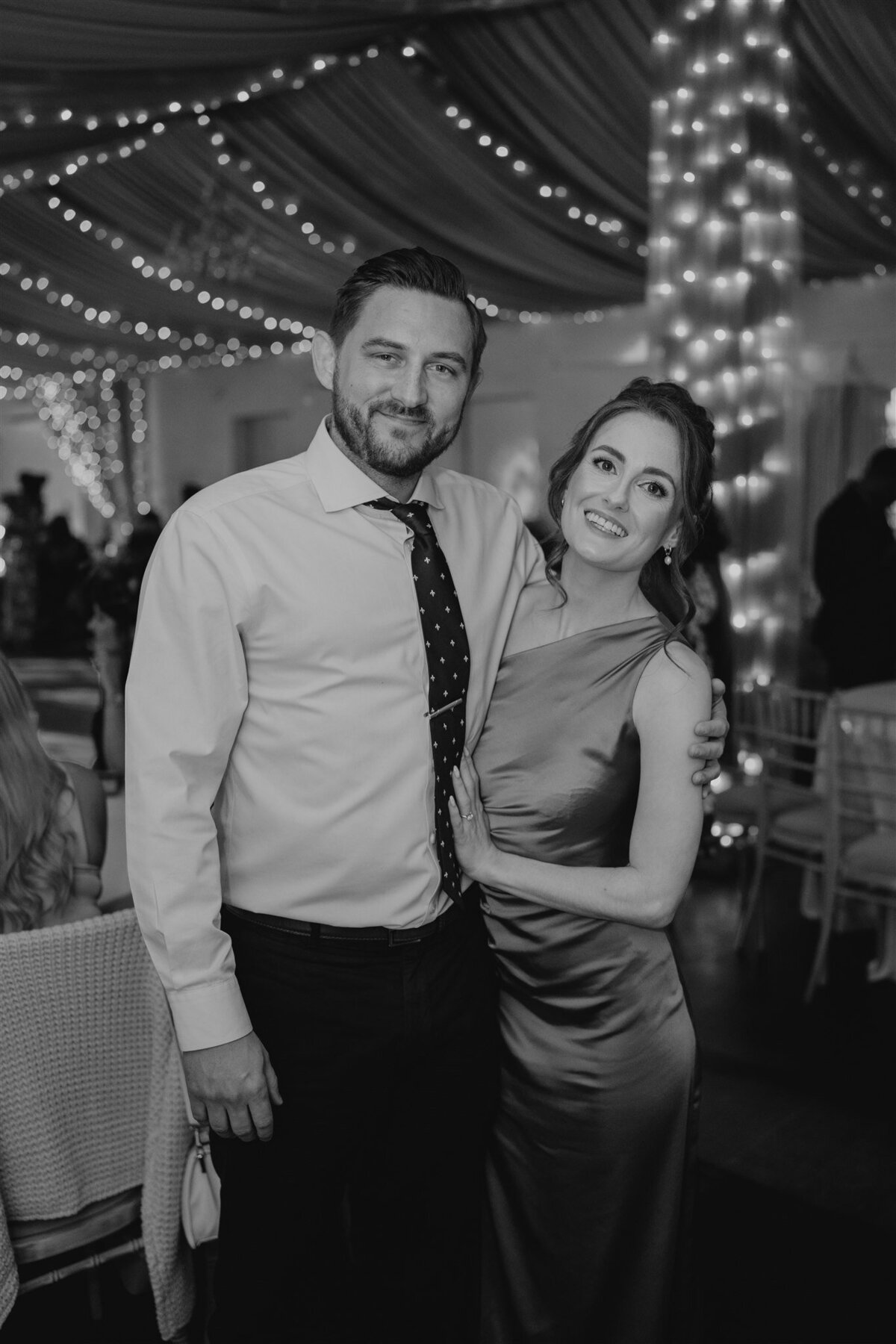 A man and women hug and pose for a photograph in a large modern ballroom at Logie County House. People dance behind them and fairy lights hand overhead.