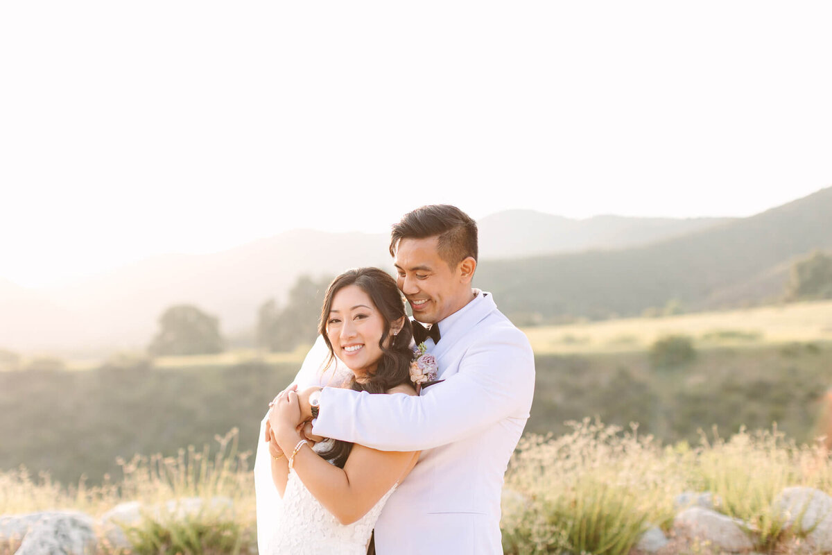 A couple embraces warmly in a scenic outdoor setting, surrounded by rolling hills and soft sunlight during golden hour at California wedding venue Serendipity Garden.