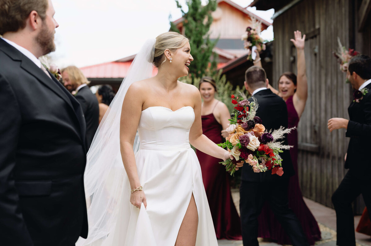 bride laughing as she walks among her bridal party who are celebrating and her groom is reaching for her hand after their outdoor ceremony