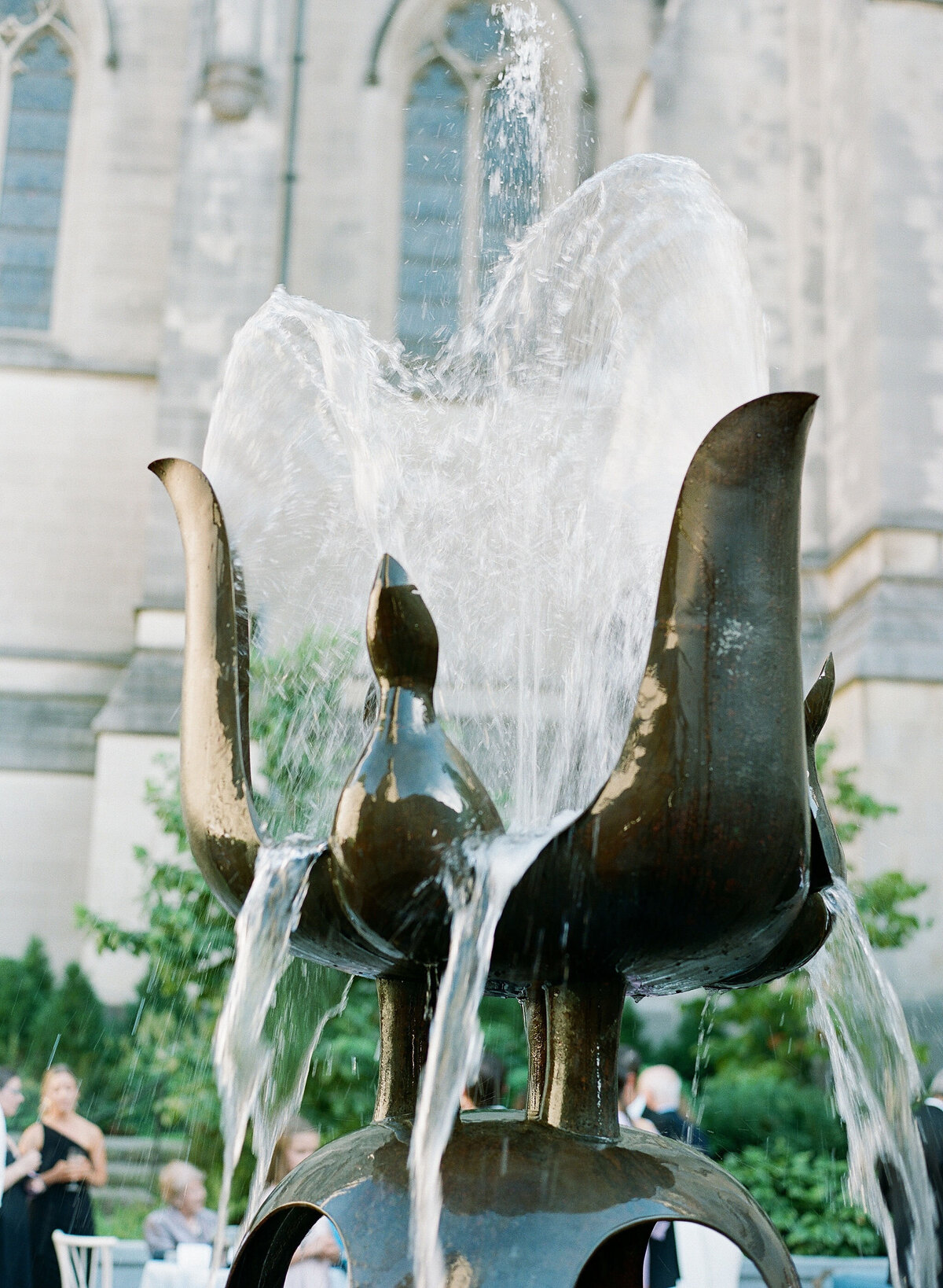 The photo was taken in Washington, D.C. at the Washington National Cathedral by photographer Twah Dougherty. In the foreground, a bronze fountain with water flowing elegantly from sculpted figures stands in front of a medieval-style stone building. People can be seen in the background, enjoying the outdoor setting. The scene is enhanced by lush greenery that surrounds the fountain, adding a sense of tranquility to the overall setting.