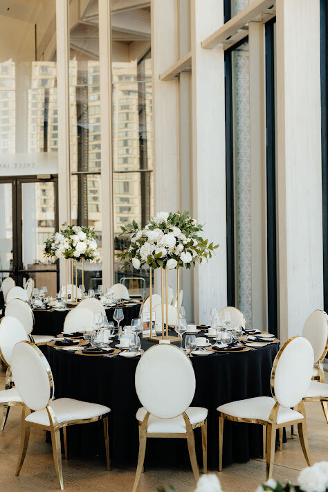 A close-up of a guest table at a National Arts Centre wedding, featuring a black satin linen with white leather chairs with gold frames surrounding the table. The centrepiece of the table is a tall floral arrangement of white and green on a gold stand, and the room is filled with natural light from the tall windows overlooking downtown Ottawa.