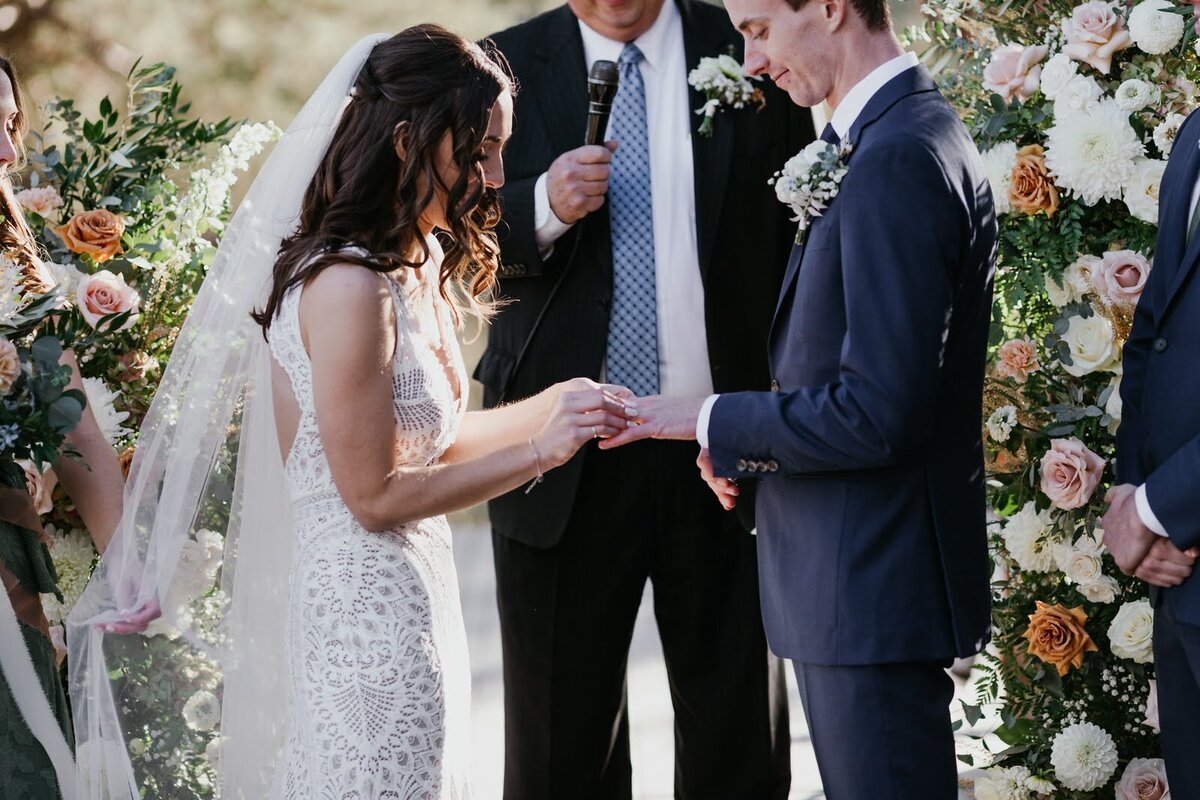 A bride and groom exchange rings and are framed by lush floral pillars by Flowers and Thyme.