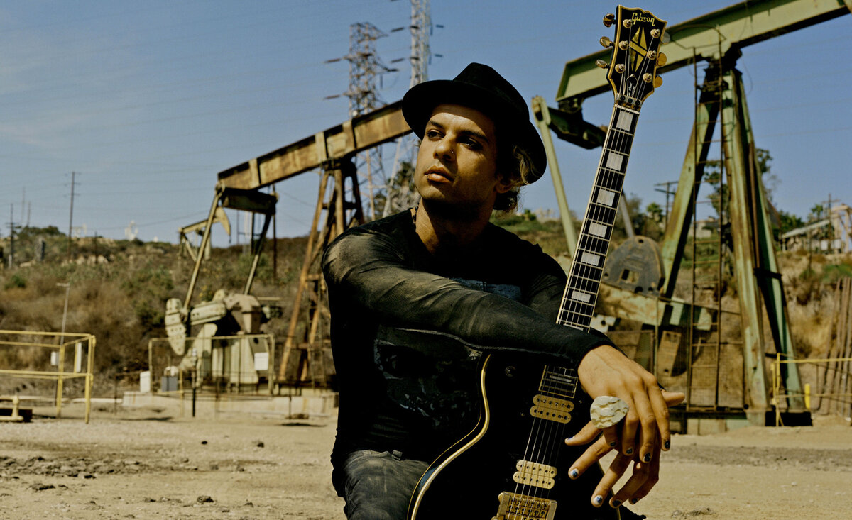 Male musician photo Fernando Basilio wearing black outfit with black fedora while kneeling holding black electric guitar with oil pumps behind
