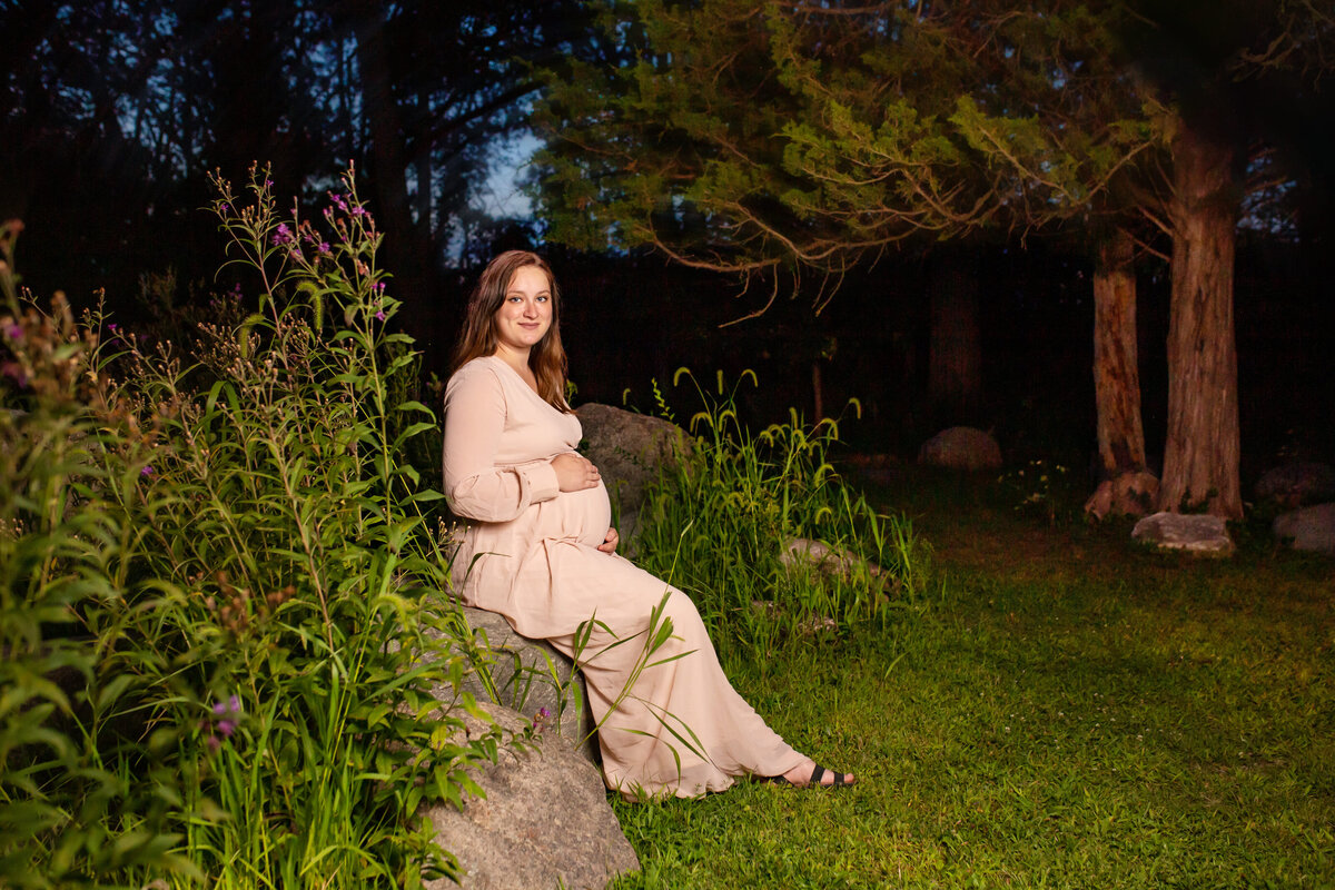 A pregnant woman sits on a rock near wildflowers and looks at the camera.