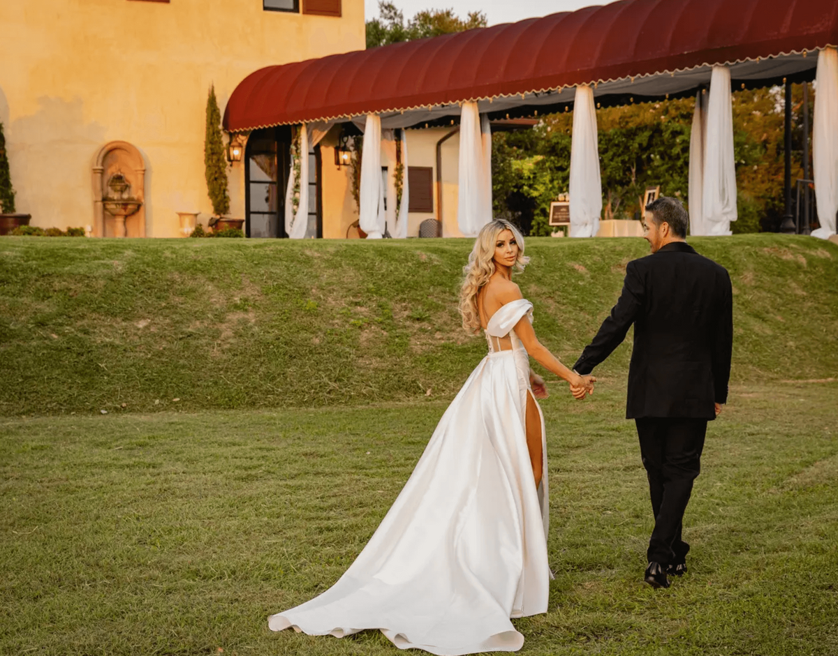 Bride and groom walk hand-in-hand at golden hour in Austin, featuring glamorous bridal waves, airbrush makeup, and a sleek satin gown with thigh-high slit