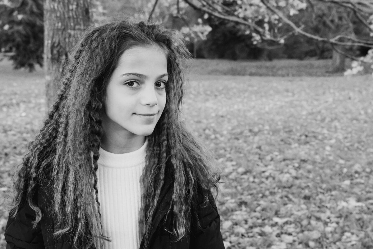 Young girl with curly hair smiling slightly, standing in a park adorned with autumn leaves, in a black and white photograph.