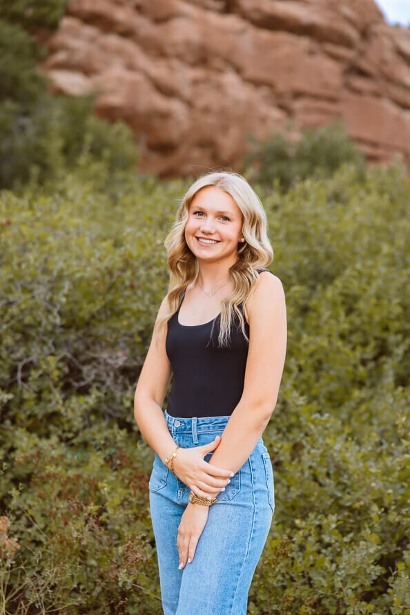 Girl holds arm and smiles with green bushes behind her.