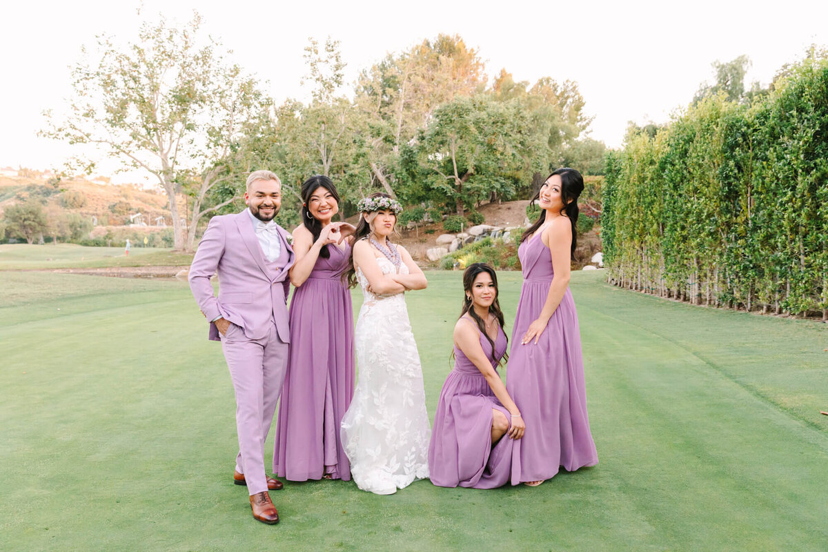 A bridal party poses outdoors on a sunny day. The bride in a white dress stands center, surrounded by attendants in lavender outfits, with greenery and trees in the background. Smiles and playful poses convey joy and celebration.