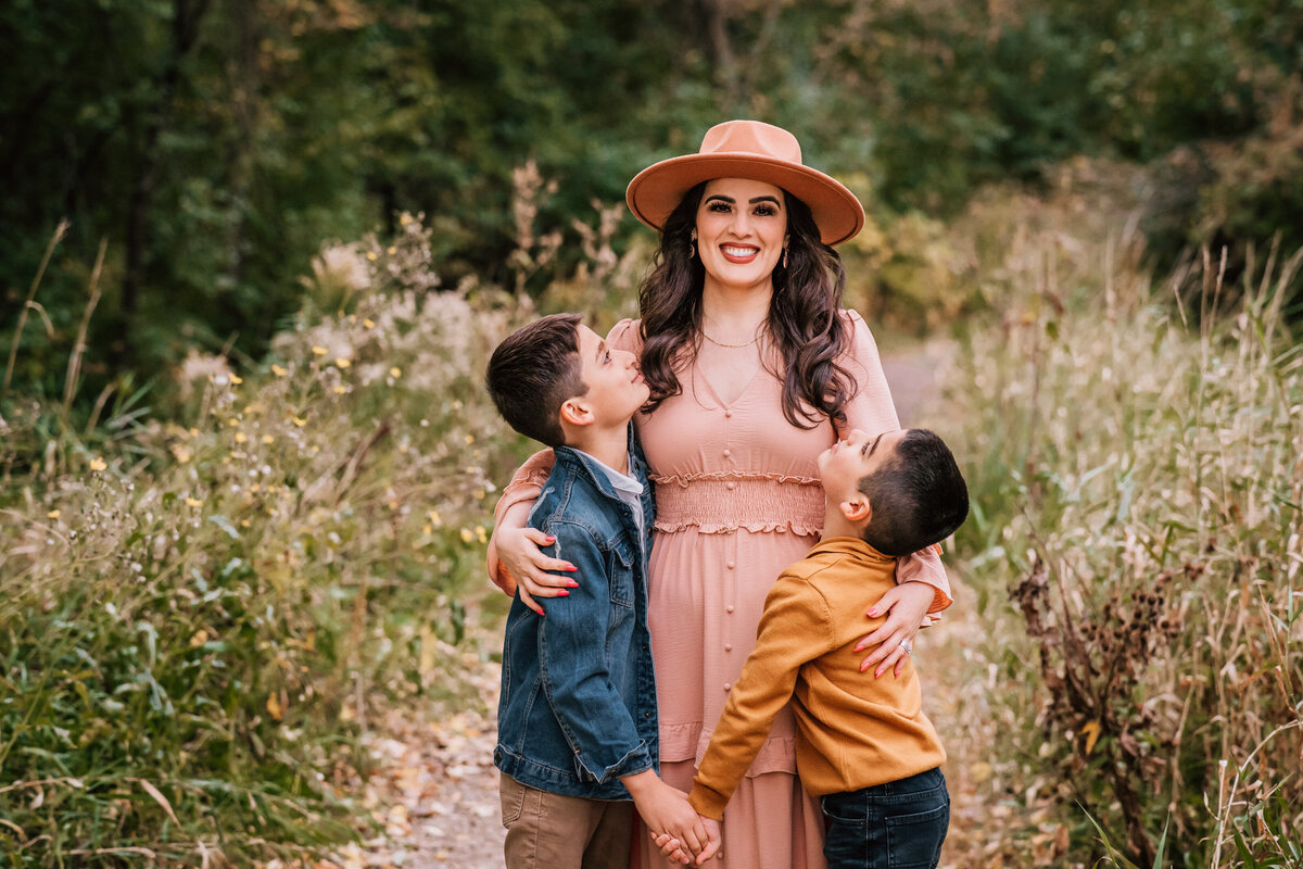 A mother, dressed in a long dress and hat, embraces her two sons, who gaze up at her with adoration. They stand together on a leafy trail surrounded by greenery.