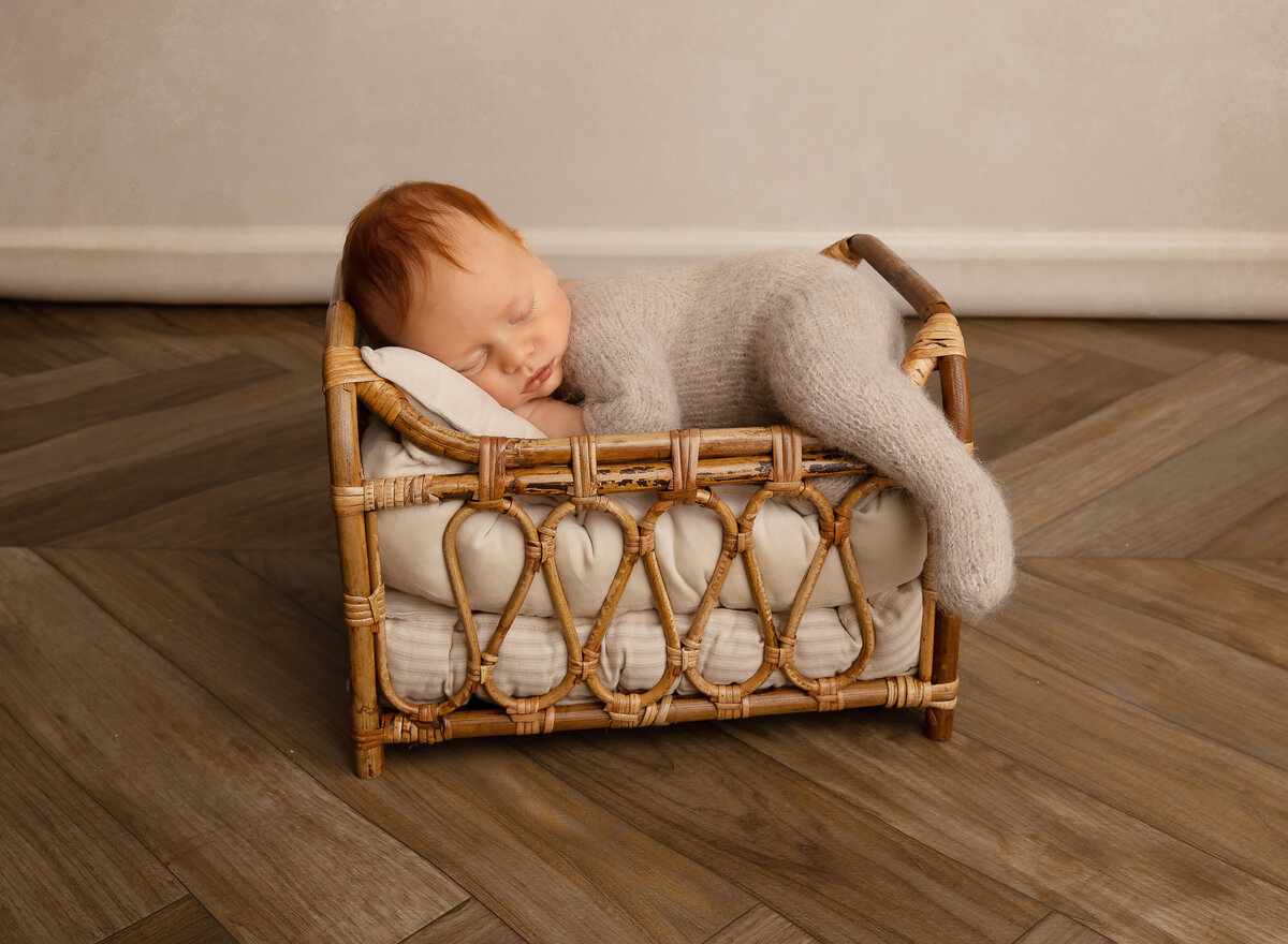 Baby boy is sleeping on his belly in a miniature bed for a newborn photoshoot. Baby's hands are placed underneath his chin. Captured by top Brooklyn newborn photographer Chaya Bornstein