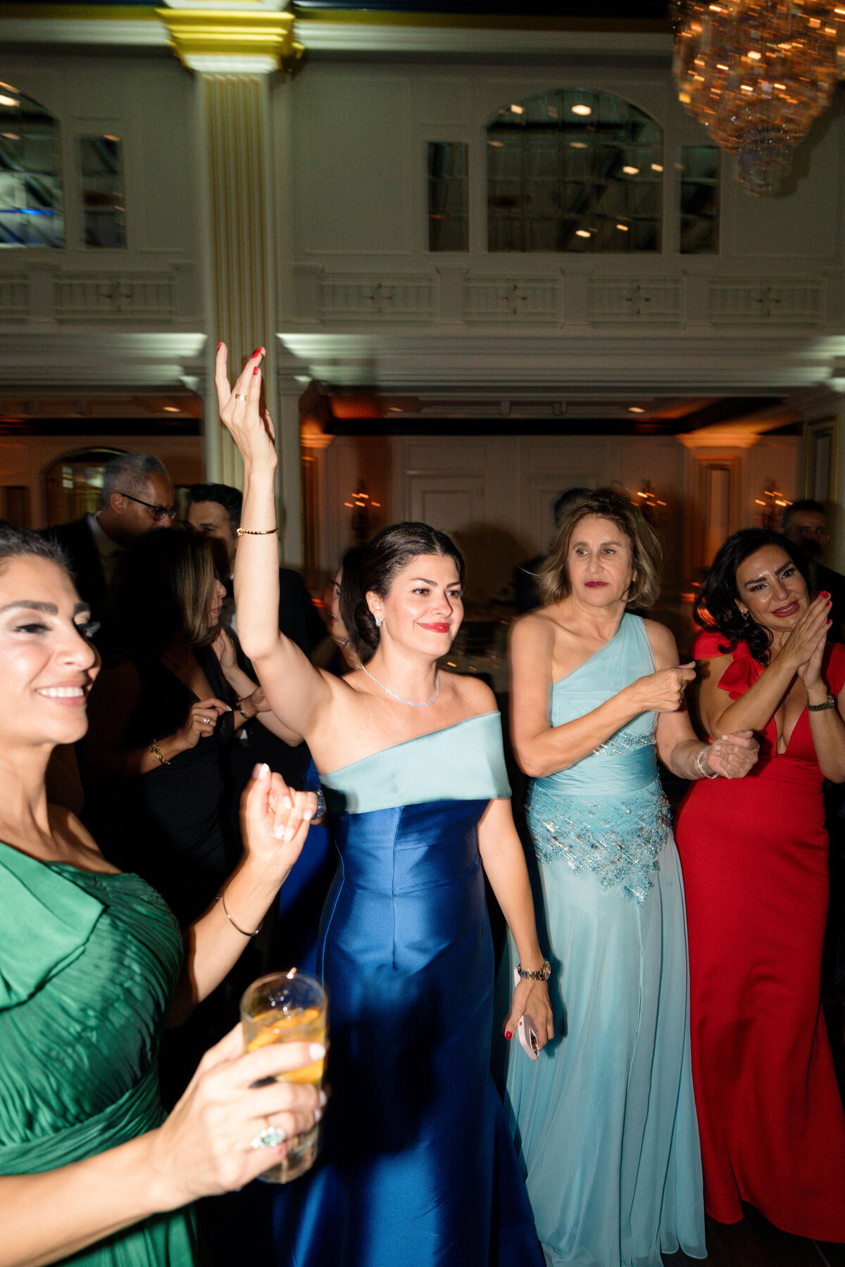 A group of elegantly dressed women dance and celebrate at a formal event. One woman in a blue gown raises her hand joyfully, while others smile and clap along. The setting is a chandelier-lit ballroom with classic decor.