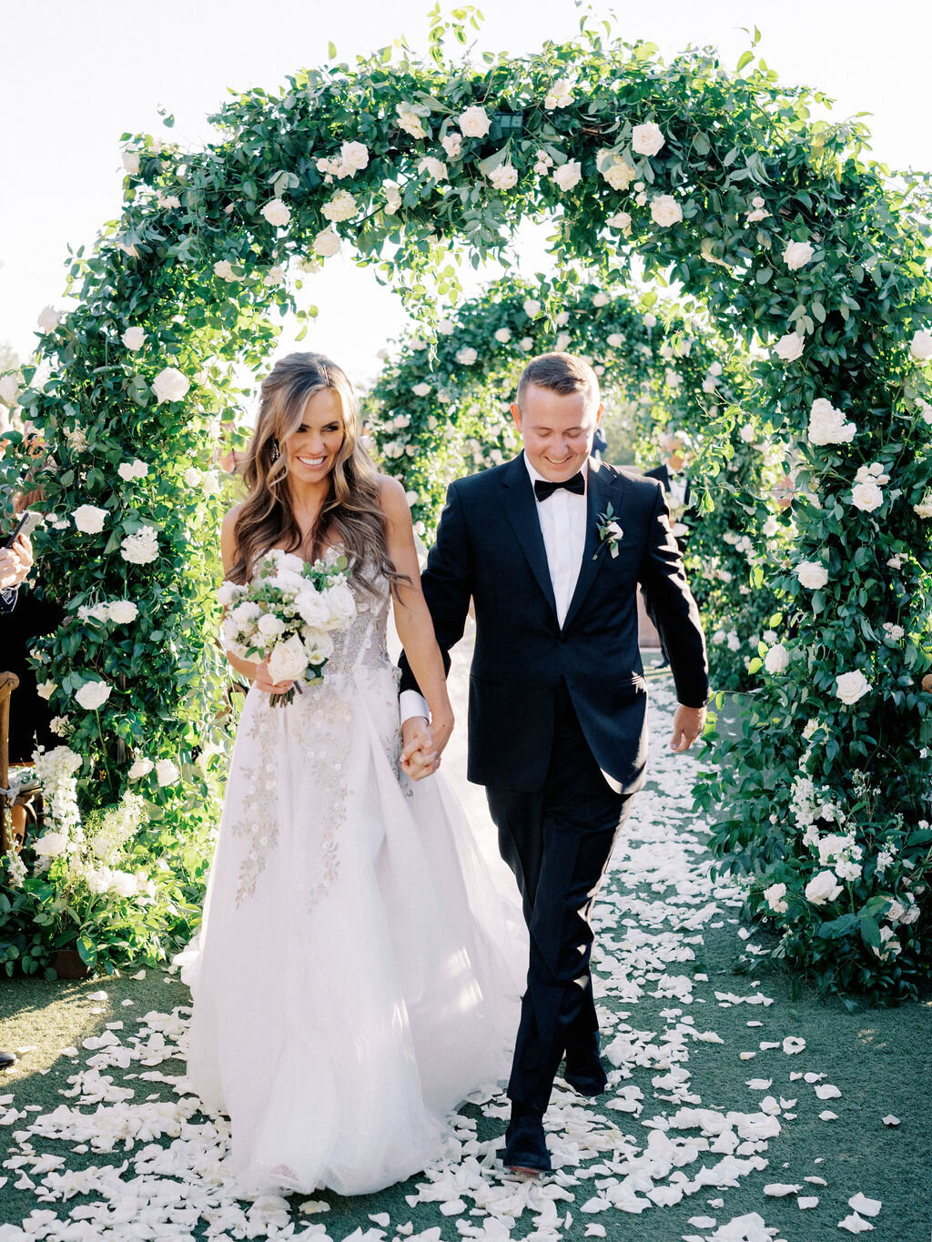 The newlyweds share a joyful moment walking hand-in-hand down the aisle under lush floral arches, surrounded by wedding guests. A romantic and unforgettable wedding celebration.