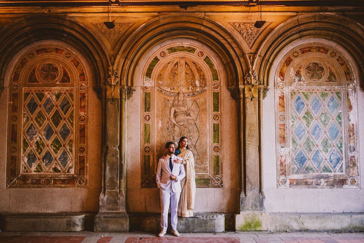 An engaged couple wearing Indian sari and suit standing in front of a mural arch inside the Bethesda Terrace in Central Park