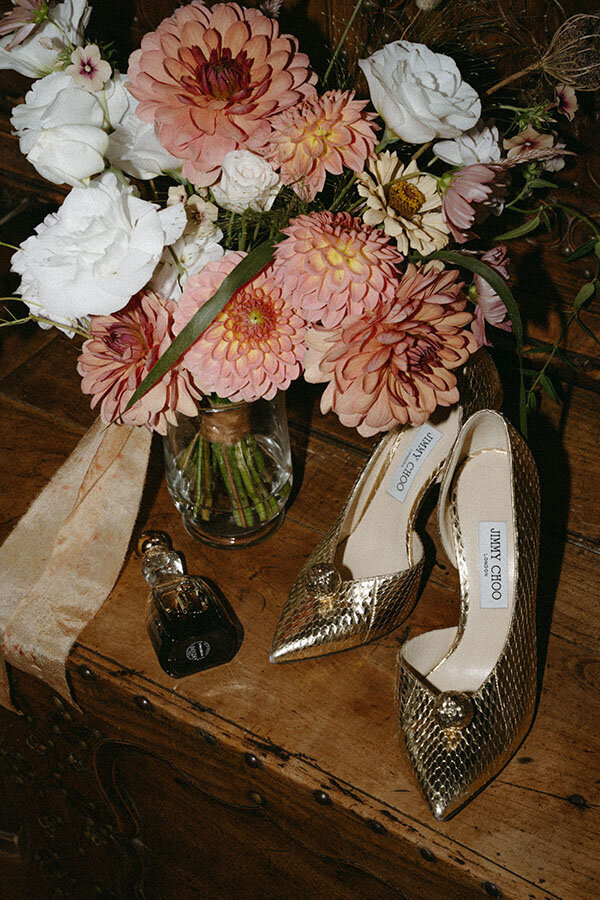 Chaussures dorées et bouquet pastel posés sur une table en bois. Mariage en bord de mer, photographié par Kamescop, photographe vintage.
