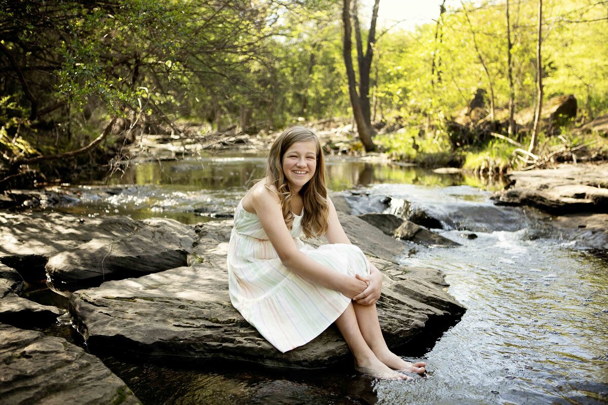 The youngest sister standing in the shallow creek in Flower Mound, smiling joyfully as the water gently flows around her