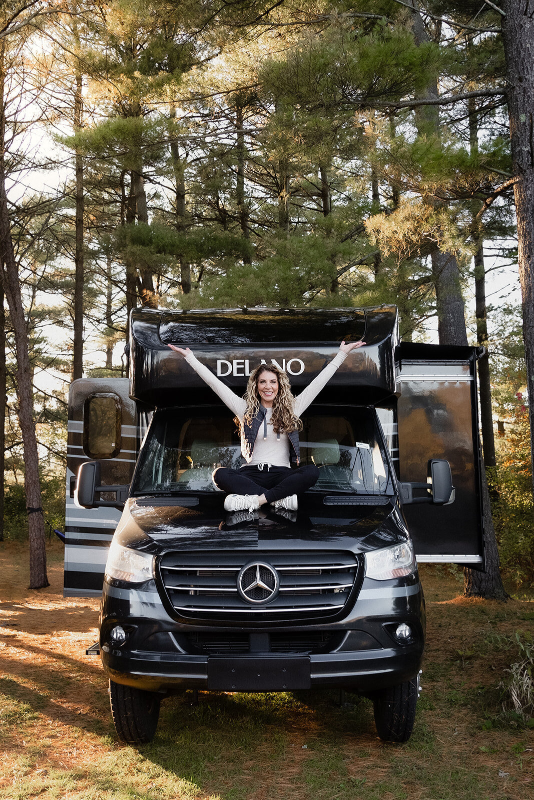 woman sits on the hood of an RV  with her hands up in the air.