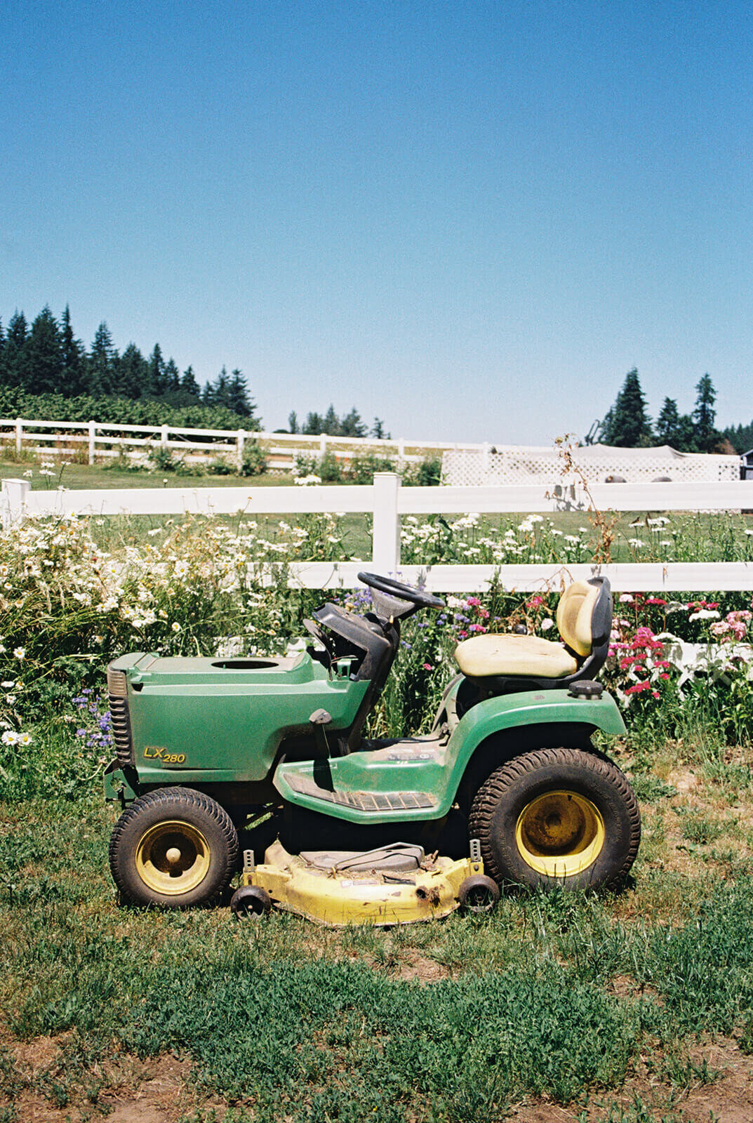 A lawn mower in front of a white picket fence and flowers.
