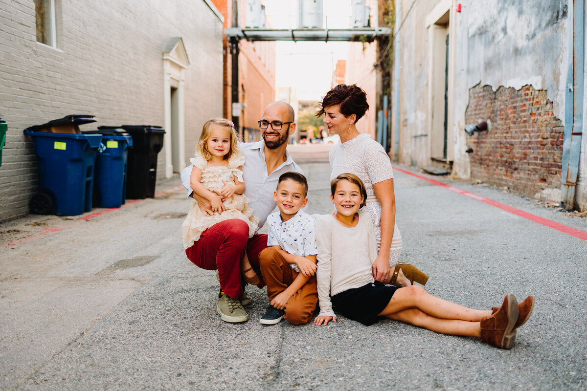 Family photo featuring a man in glasses and a blue shirt sitting on the street while observing a girl holding onto his leg. The blonde girl is looking into the camera, with two boys sitting on the floor nearby