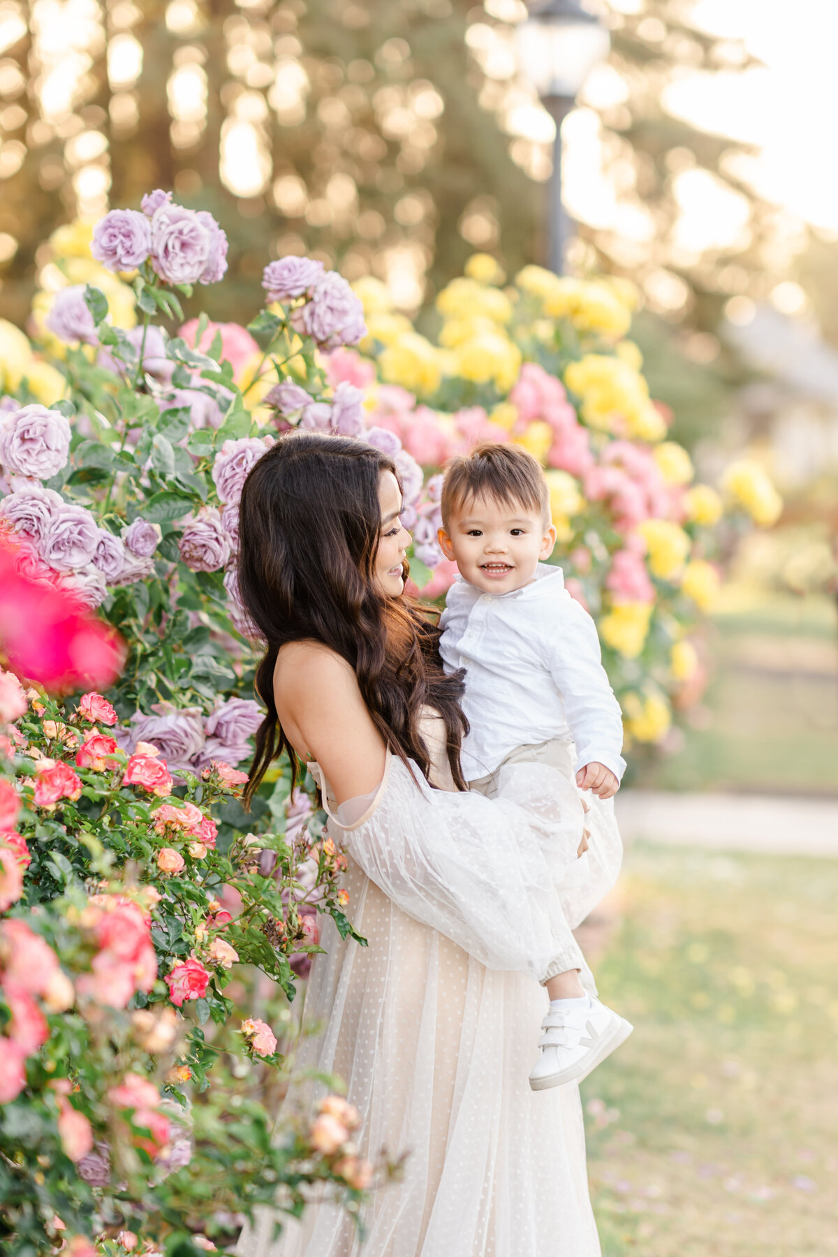 A mother is holding and smiling at her son while standing in a rose garden as the little boy smiles photographed by Bay area photographer, Light Livin Photography