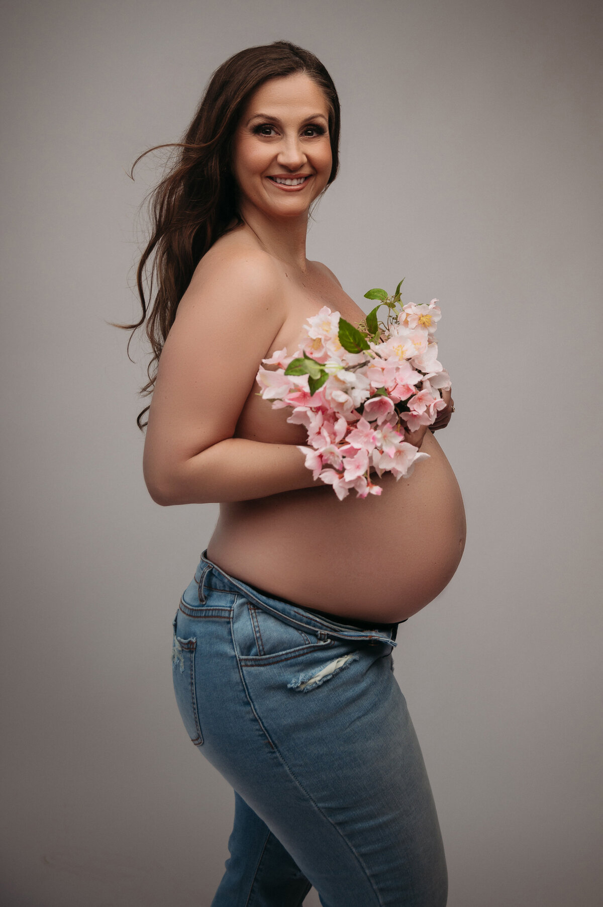 Pregnant brunette woman holding pink  flowers over her baby bump and wearing jeans in front of grey background