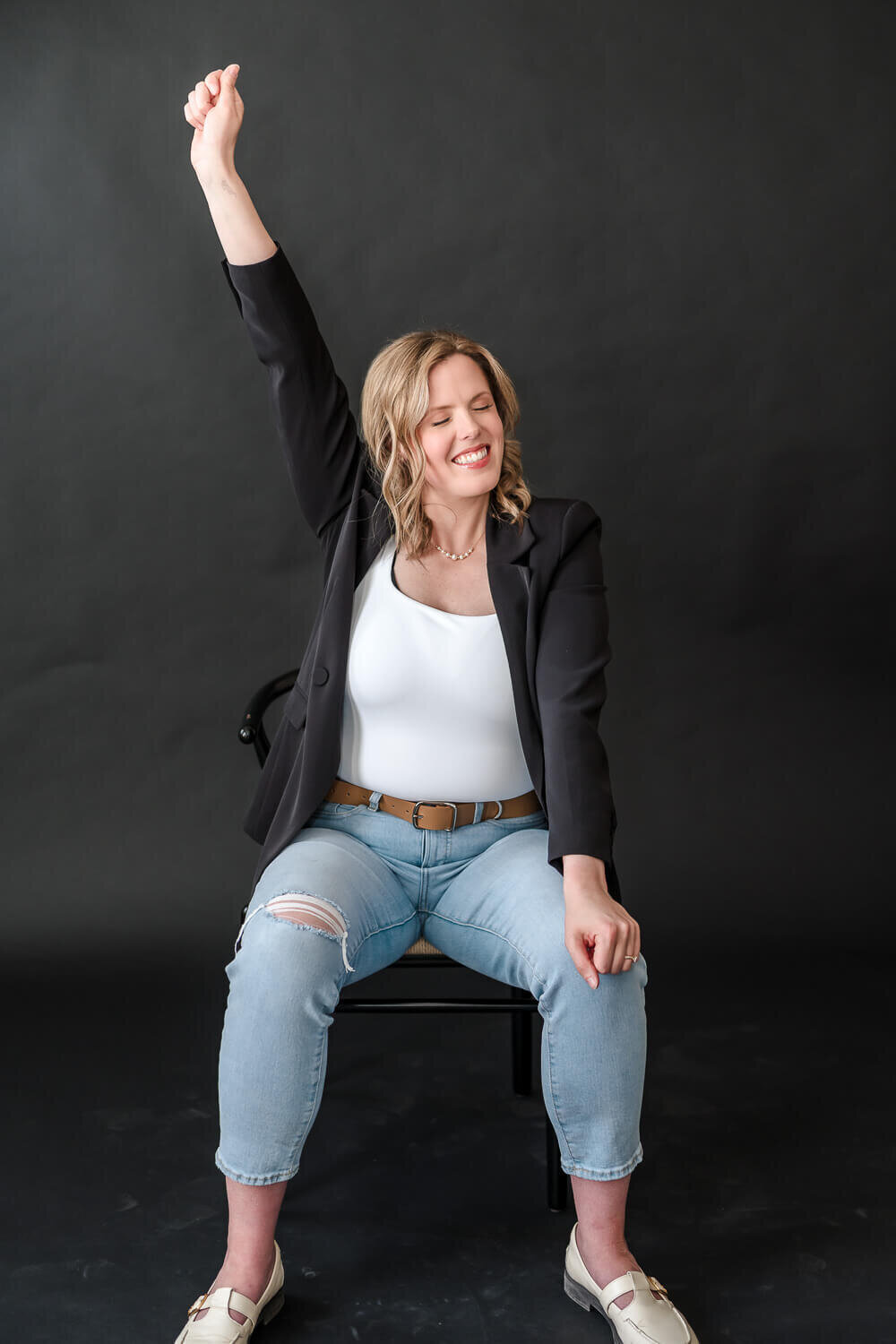Joyful female entrepreneur sitting on chair with arm raised in celebration, eyes closed, smiling.