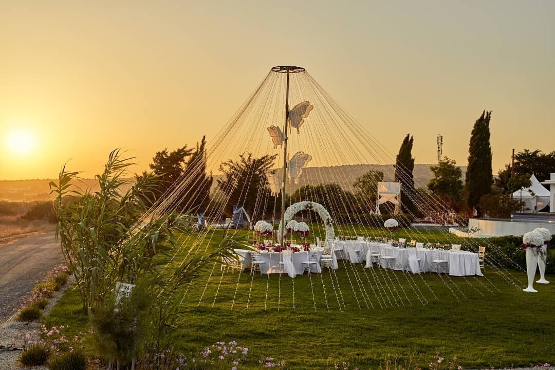 A tall fairy light curtain reaches high into the sky surounding a banqueting table at sunset