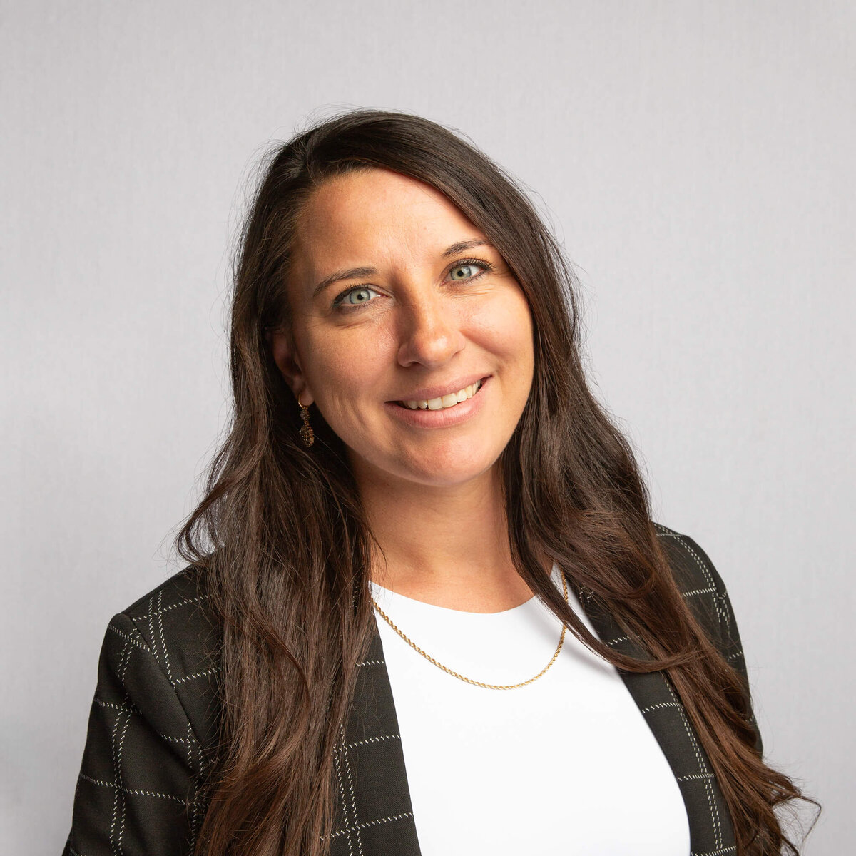 Headshot photograph of a woman with dark brown hair, a white top, and a dark jacket, in front of a light grey background.