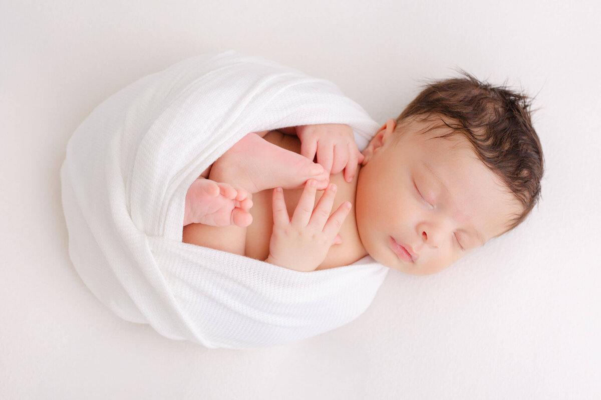 tiny newborn baby sleeping peacefully while wrapped up in a white swaddle with feet and hands showing