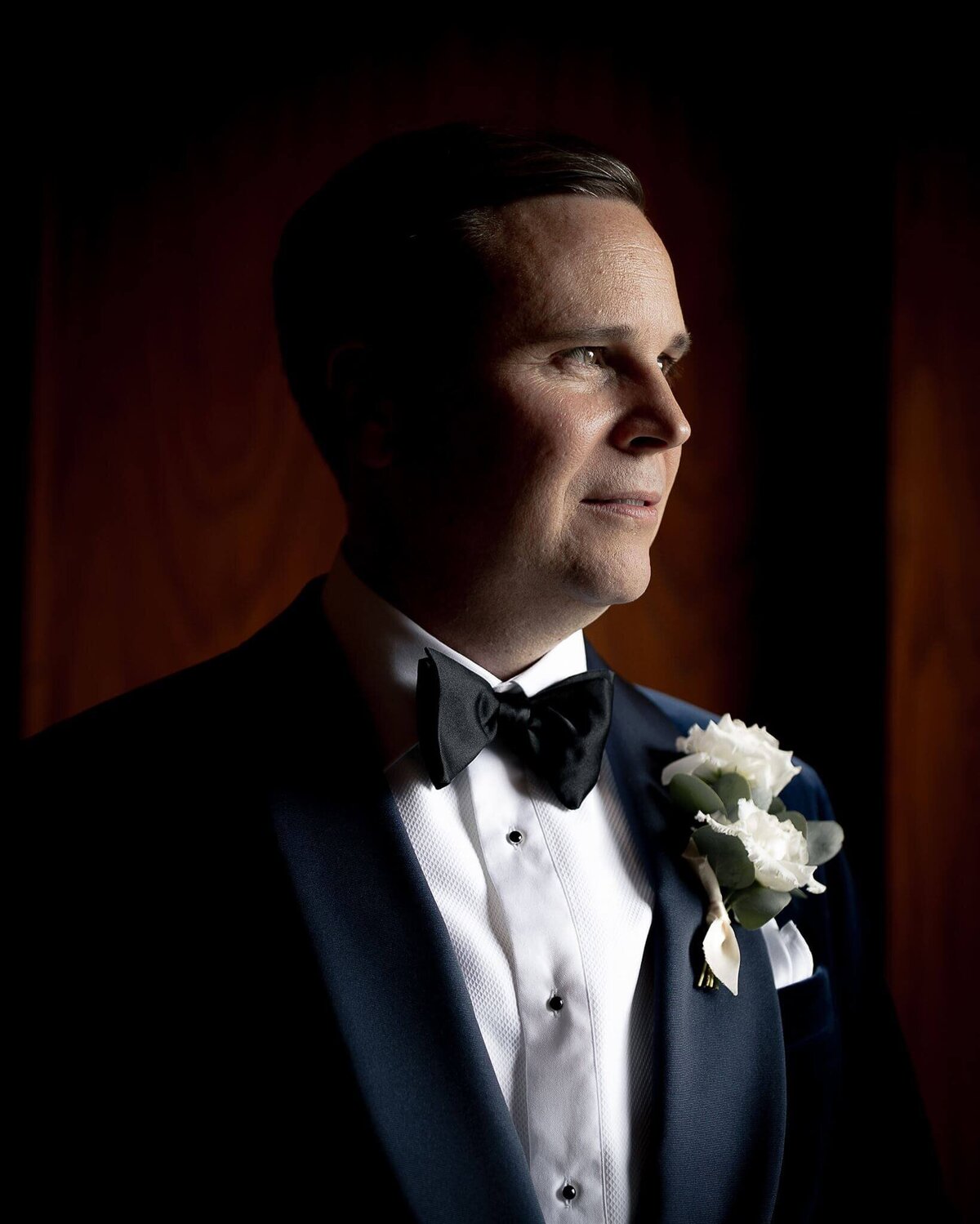 A groom looks to the right wearing a blue velvet jacket and white flowers