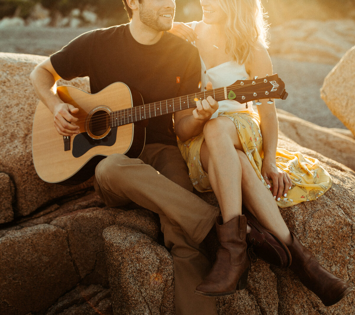 A girl in a yellow skirt is sitting on a rock next to her fiancé while he plays the acoustic guitar with the sunset behind them,
