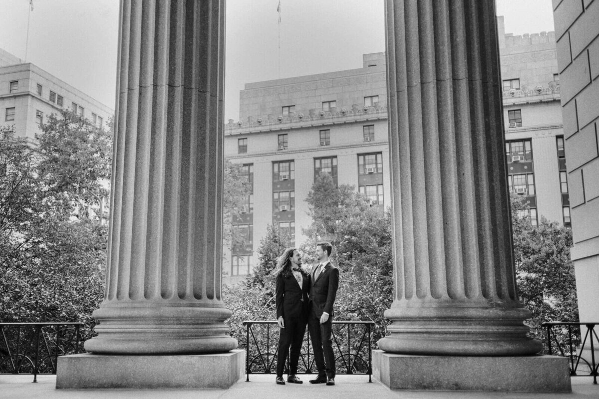 The two grooms are closely standing in between two large columns of the NYC City Hall. Elopement Image by Jenny Fu Studio
