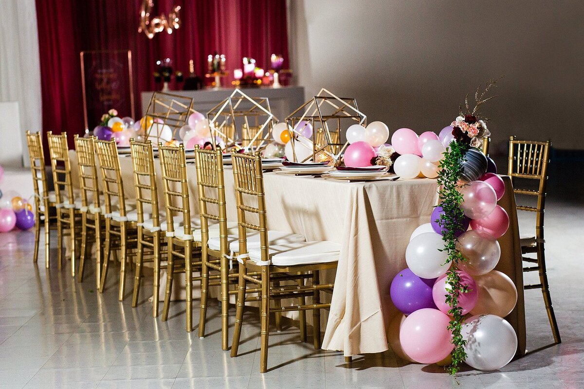Long table with an ivory tablecloth and gold Chivari chairs set with a long garland of pink, white, peach and purple balloons and gold geometric cubes.