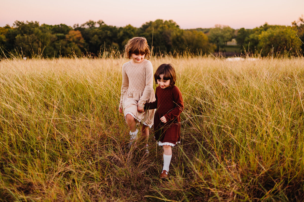 Photo of two sister girls in a garden with high grass, they are holding hands. The youngest has a red white dress and is facing the floor. The other girl has a beige dress