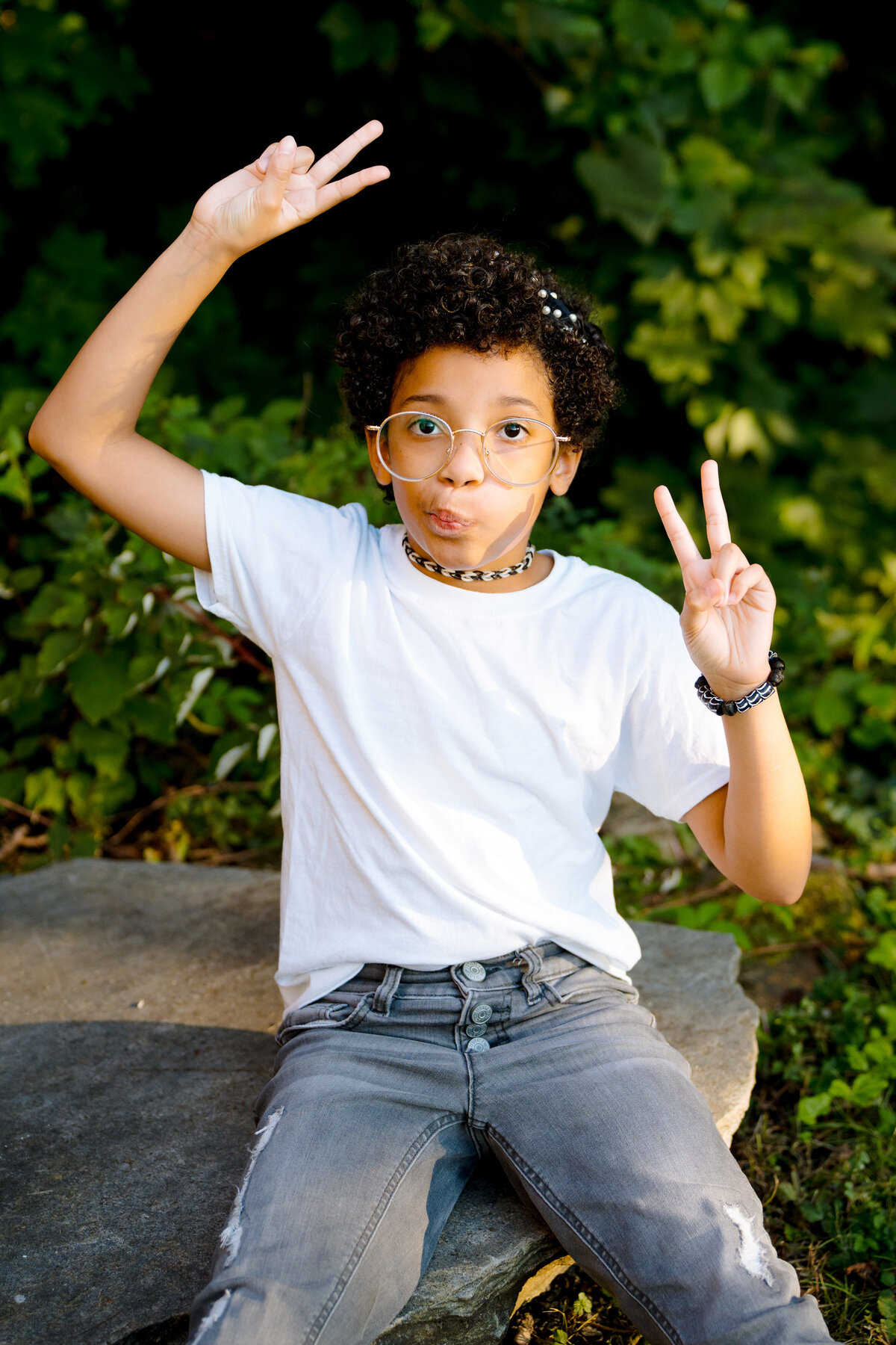 A young child making a funny face and holding up peace signs while sitting on a rock.