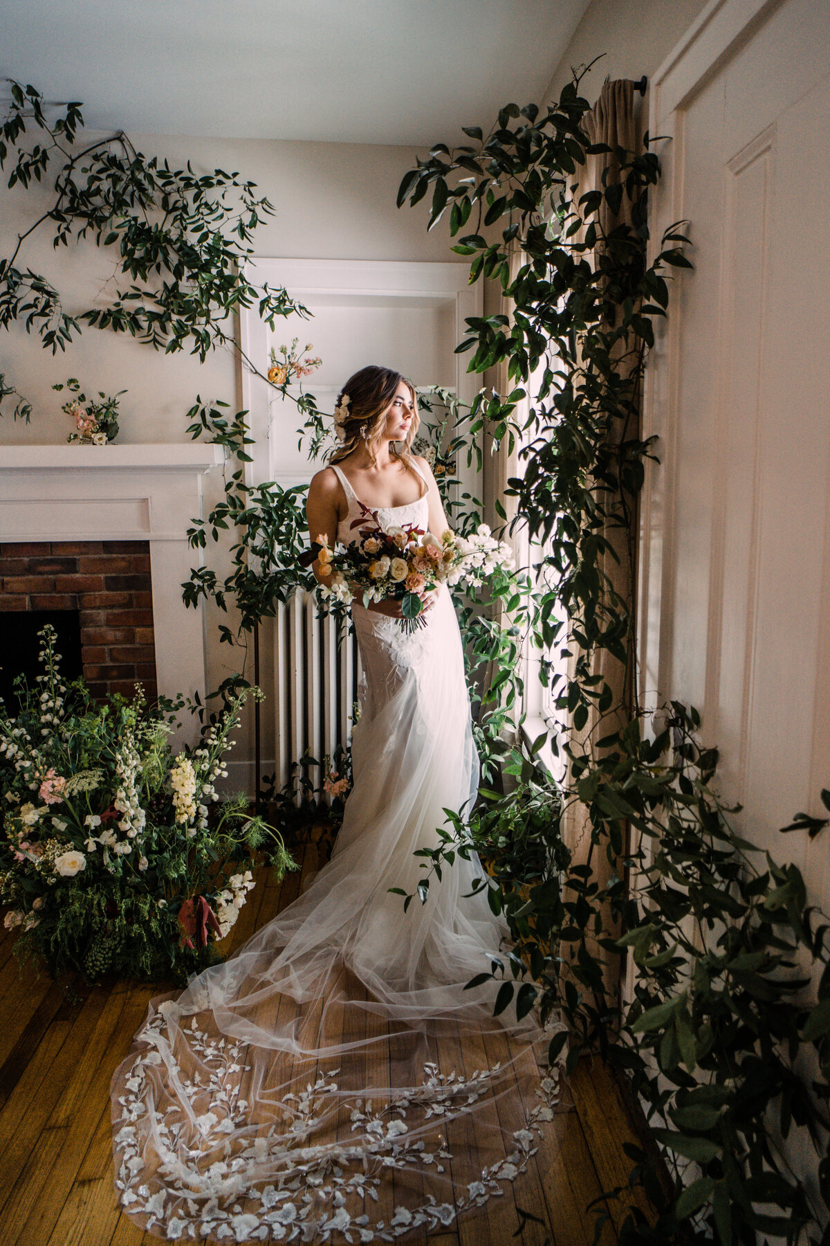 Bride wearing Alexandra Grecco lace gown & floral applique veil holding bouquet in indoor flower garden with vines on walls