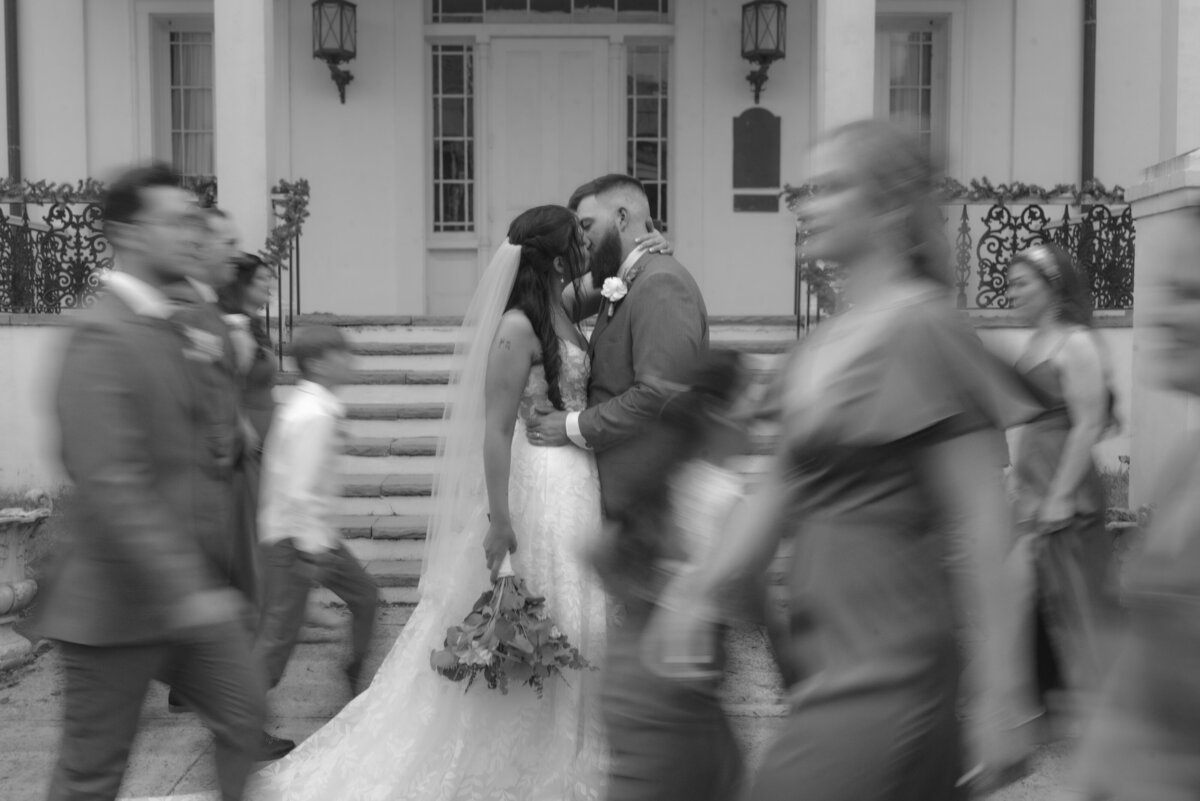 a bride and groom kissing while their wedding party walks around them adding some motion blur to the photo.