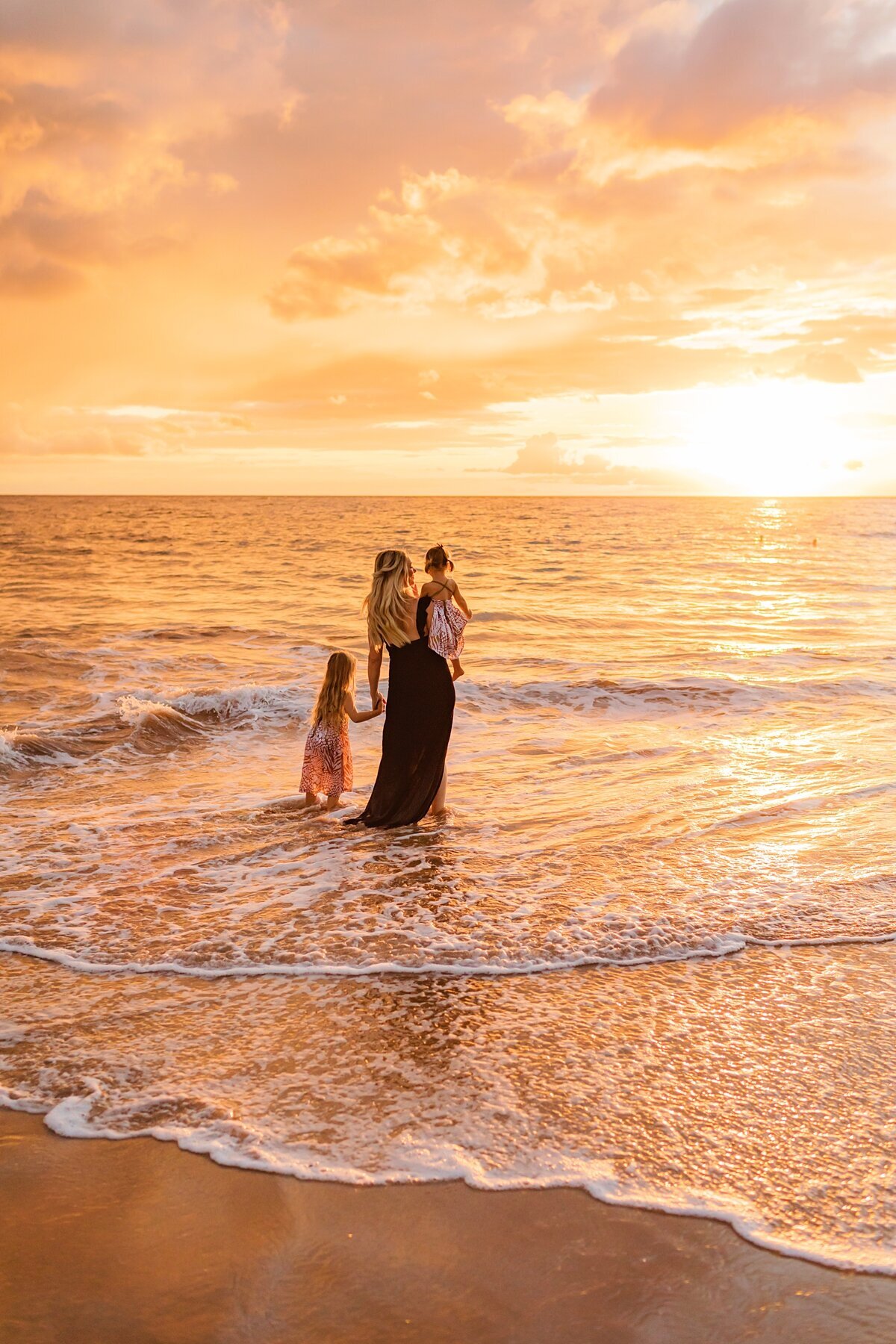 Mother wearing dark blue dress holds her daughters next to the ocean for their sunset family photoshoot at the beach on Maui