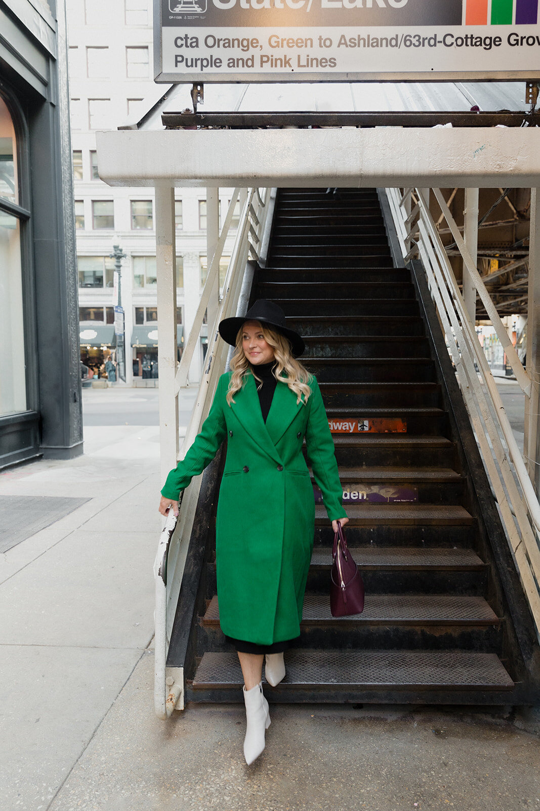 blonde woman is walking down the steps of the Chicago L stop in a green coat and black hat. She carries a  purse and looks off to the side.