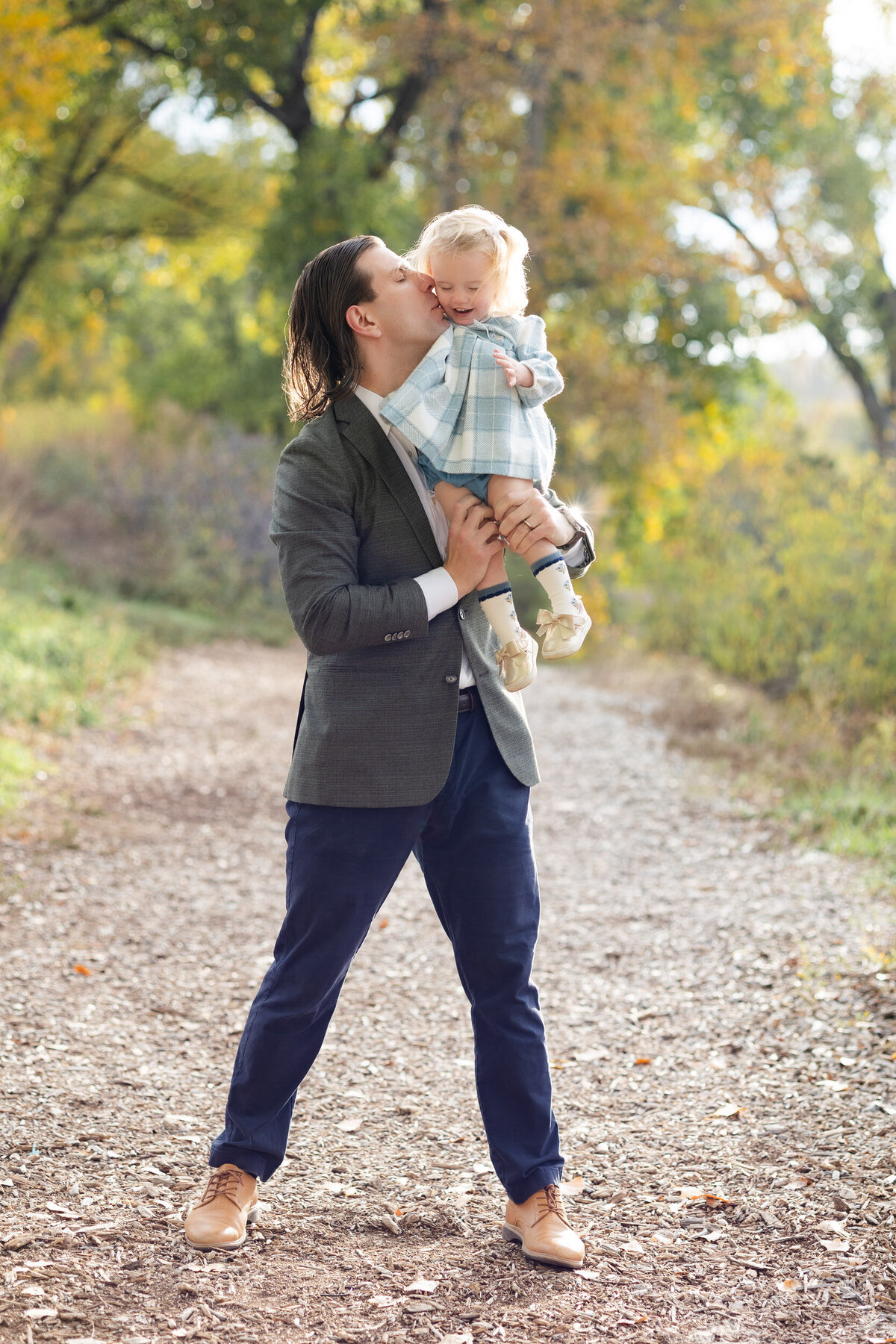 A dad holds his toddler daughter on his shoulder and kisses her cheek.