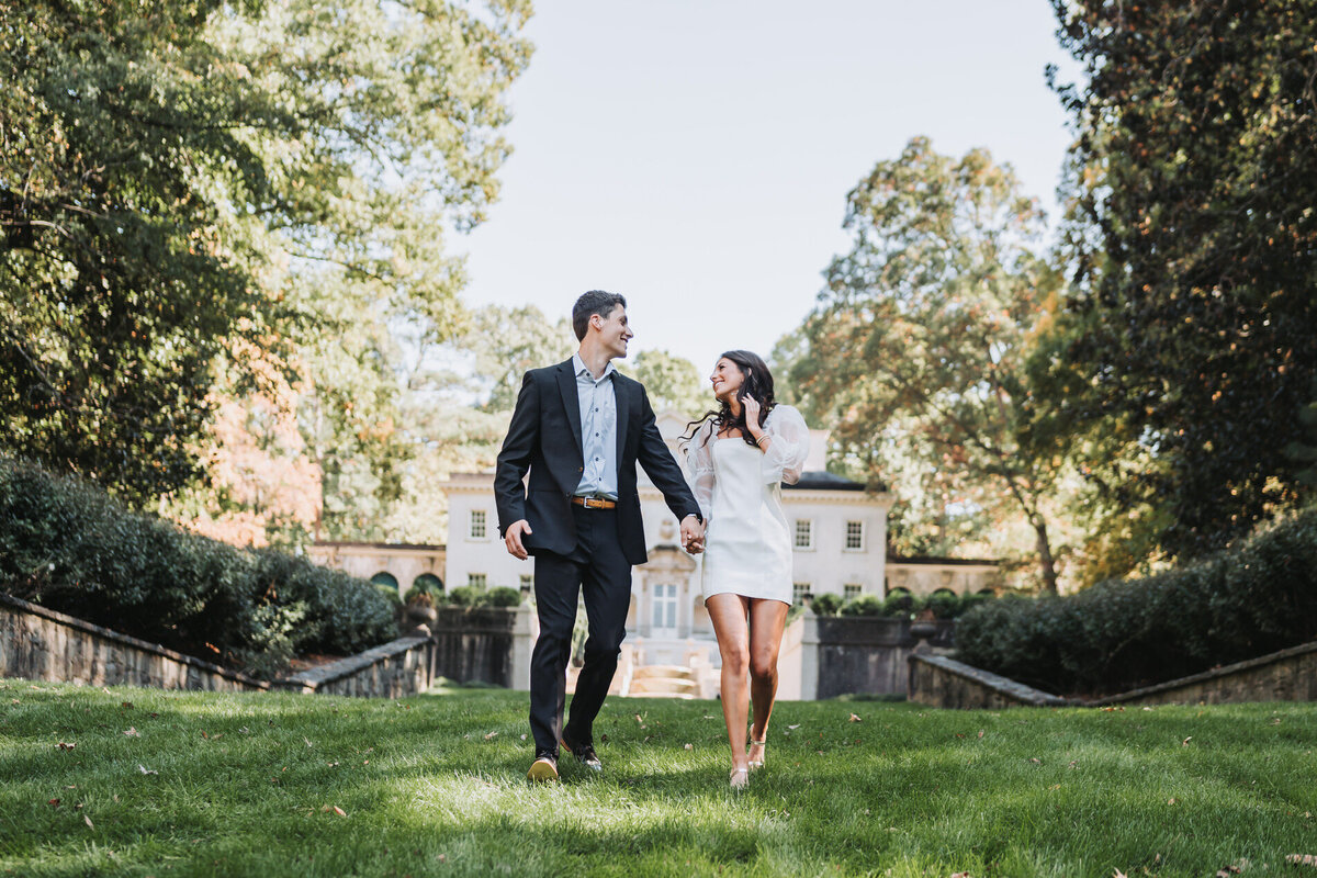 couple holds hands and run towards camera with swan house in the background