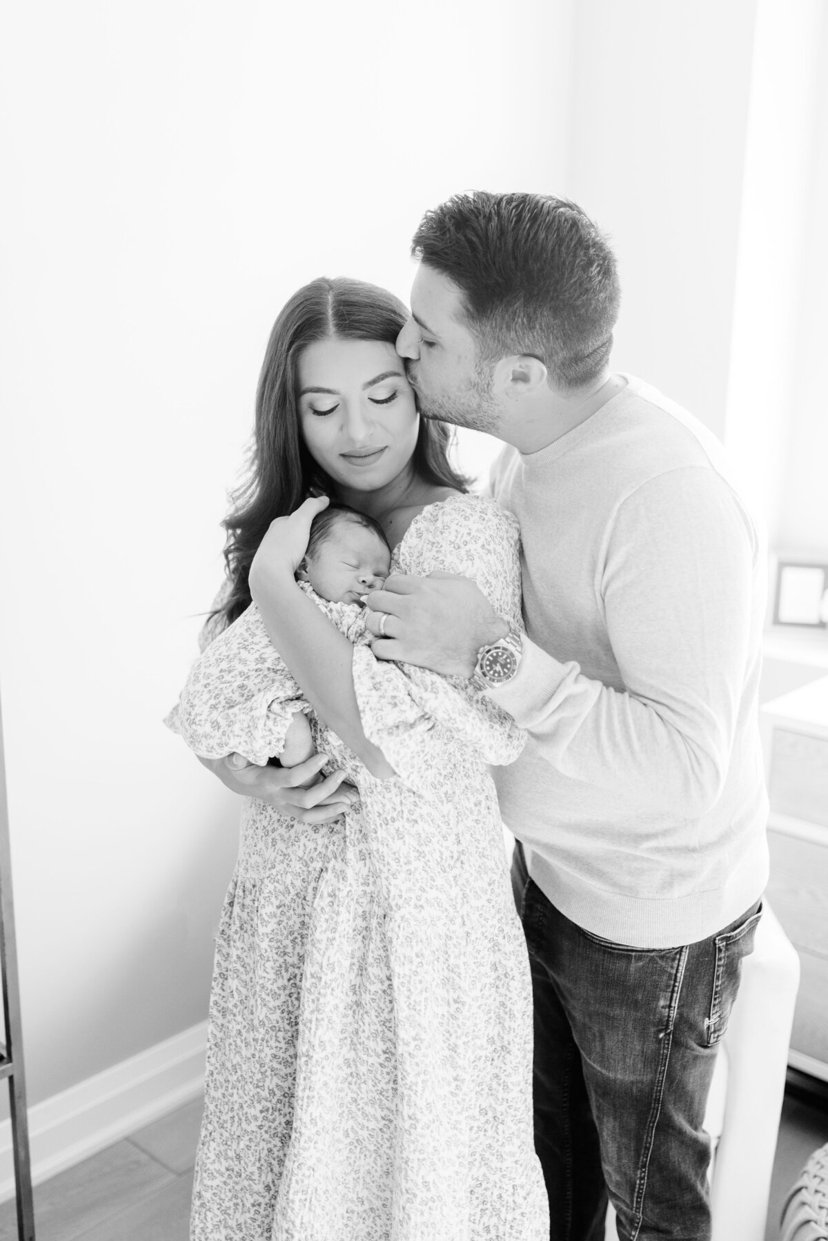 Couple standing in nursery holding their newborn, father kissing mother’s temple – peaceful family connection