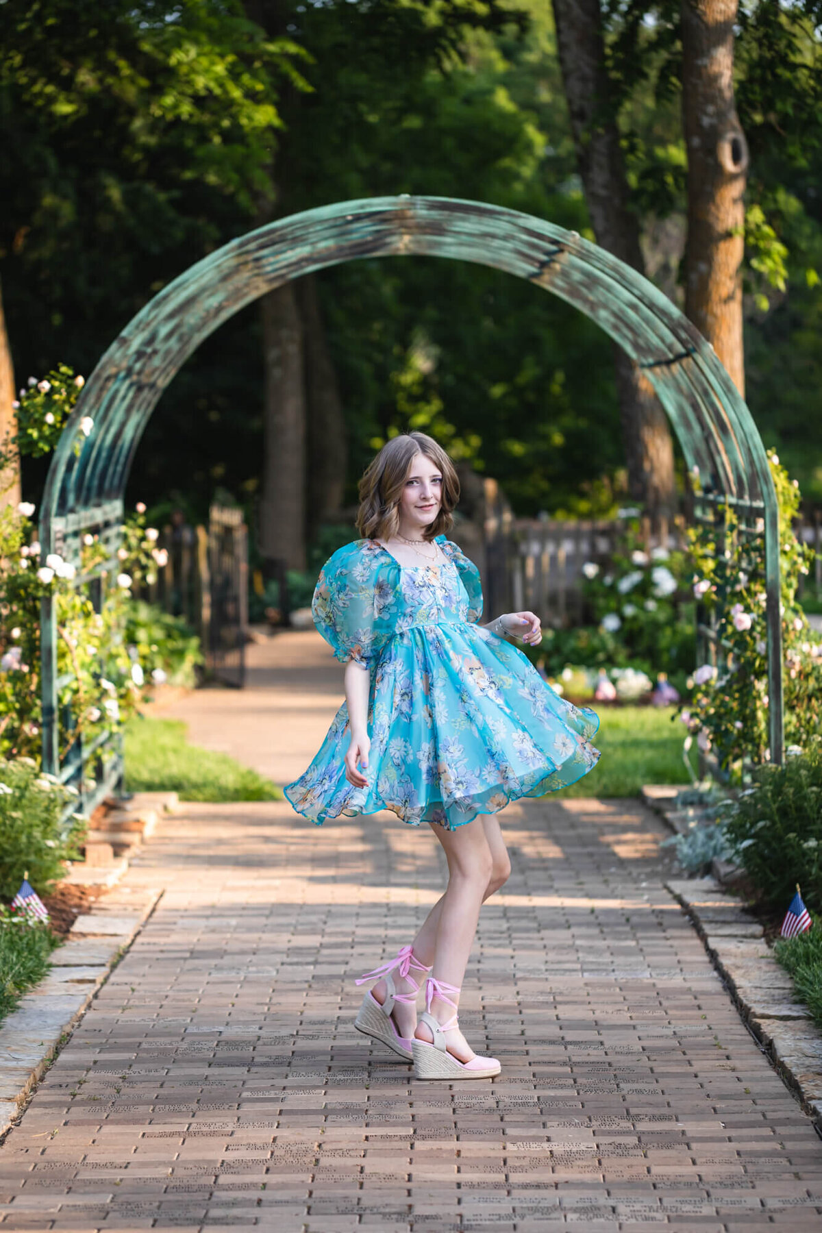 Pretty teenage girl in a blue floral dress and pink shoes twirling in front of a rose trellis. Captured by Springfield, MO teen photographer Dynae Levingston.