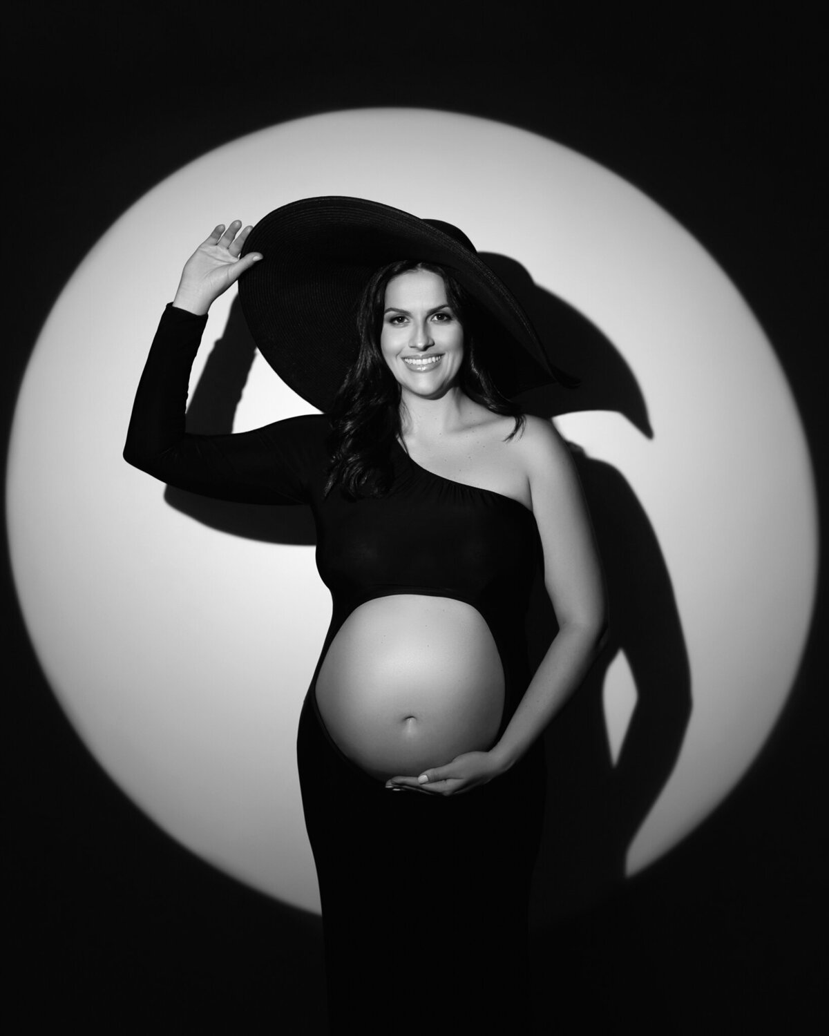 Studio maternity portrait in spotlight with one-shoulder black gown and wide-brim hat, taken in Austin, Texas
