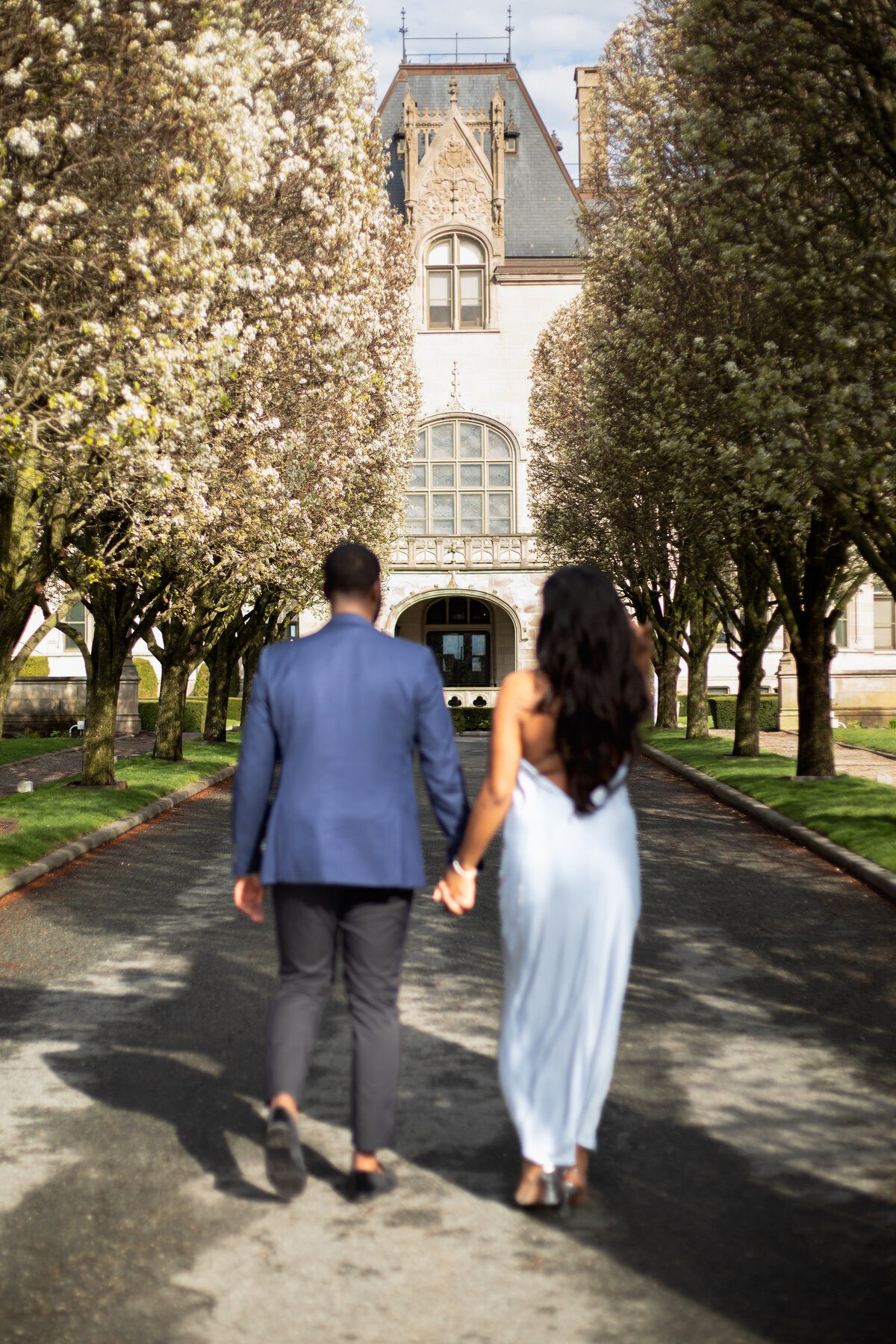 Ochre Court Mansion  | Kelsey Sheehan Photography Timeless Rhode Island Weddings | A couple holds hands, walking down a tree-lined path toward a grand building. The scene is serene, with blooming trees and a romantic atmosphere.