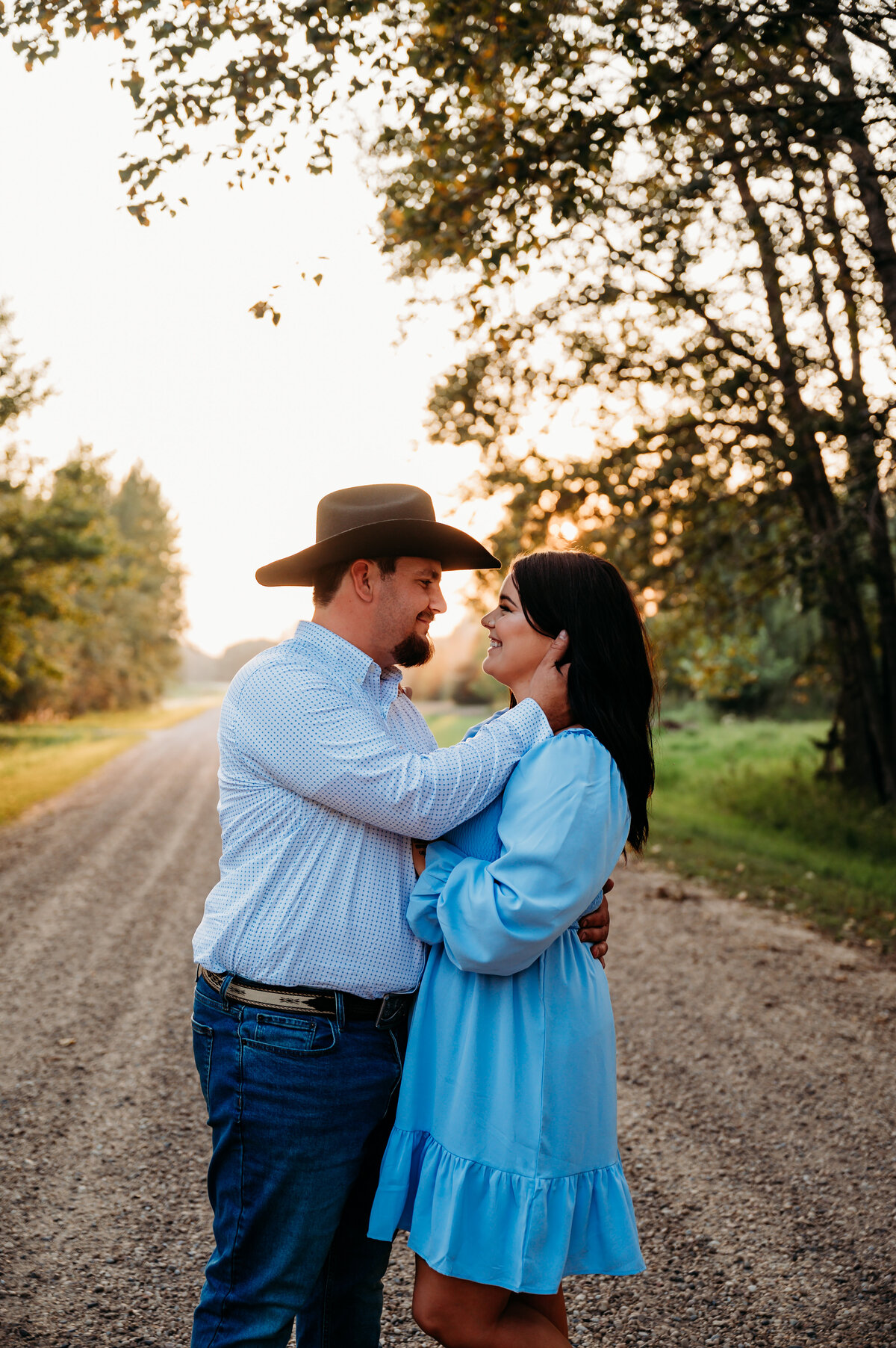 surrounded by green trees, a beautiful dirt road and golden hour sun an engaged couple look at each other in love